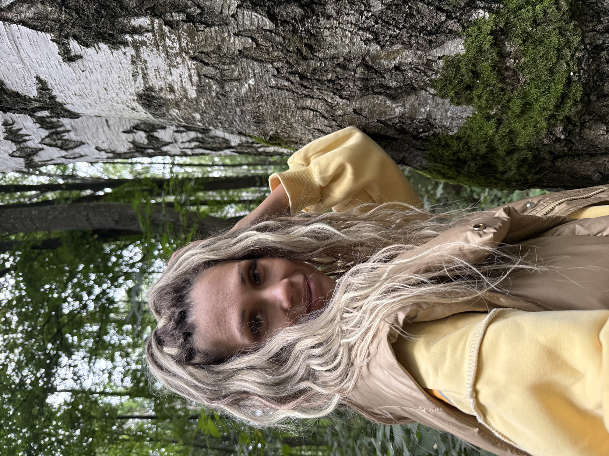 A woman with blonde, wavy hair is standing outdoors, leaning against a birch tree. She is wearing a yellow sweatshirt and a tan vest. The background is a forest with trees and greenery. There is moss growing on the birch tree. The image appears to be a selfie taken by the woman. The location is specified as Rusanovo, Russia.