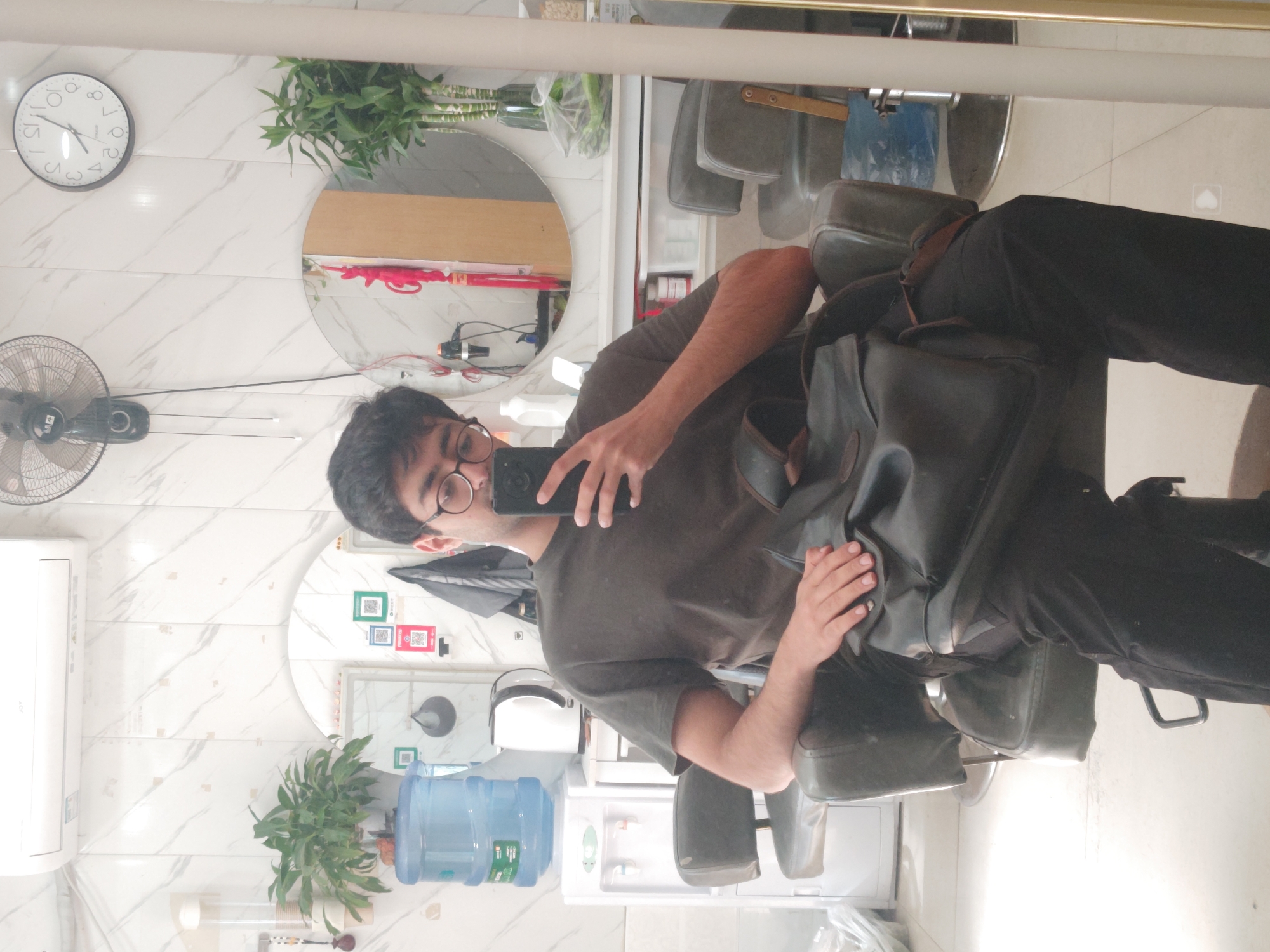 A young man with glasses and dark hair sits in a dark chair, holding a black smartphone to take a selfie. He is wearing a dark t-shirt and black pants, and a dark leather backpack rests on his lap. The background appears to be a hair salon or barber shop, with white marble-patterned walls, a water cooler, a white electric kettle, and various plants. A round mirror reflects a portion of the room, including a black fan mounted on the wall and a clock showing approximately 1:35. The overall lighting is bright, suggesting an indoor environment. This scene is located in the Chongqing High-tech Zone, China.