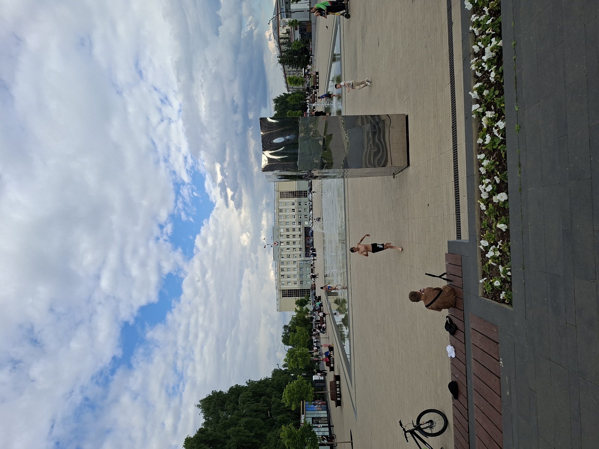 The image depicts a contemporary urban plaza in Izhevsk, Russia, with a focus on public space and leisure. The scene is characterized by a modern, reflective sculpture that dominates the foreground, catching the sky and surrounding architecture in its polished surface.
Several people are engaged in various activities. In the mid-ground, a young person appears to be playing in a shallow water feature, possibly a fountain, suggesting a warm day and recreational use of the space. Another individual, a child, is walking across the paved area. Further back, other people are seated on benches, enjoying the outdoors, with some walking and interacting. A bicycle is parked in the lower right corner.
The setting is a well-maintained plaza with a tiled pavement and flower beds lining some edges, showcasing white flowers. Behind the plaza, a modern, multi-story building stands, likely an administrative or commercial structure, with numerous windows. Lush green trees form a backdrop along one side of the plaza, contributing to a pleasant, park-like atmosphere.
The sky is filled with a mix of white clouds and patches of blue, indicating partly cloudy weather. The overall lighting suggests it is daytime, likely afternoon, given the clarity and brightness of the scene. The mood appears relaxed and leisurely, with people enjoying the public amenities. No discernible text is visible in the image.