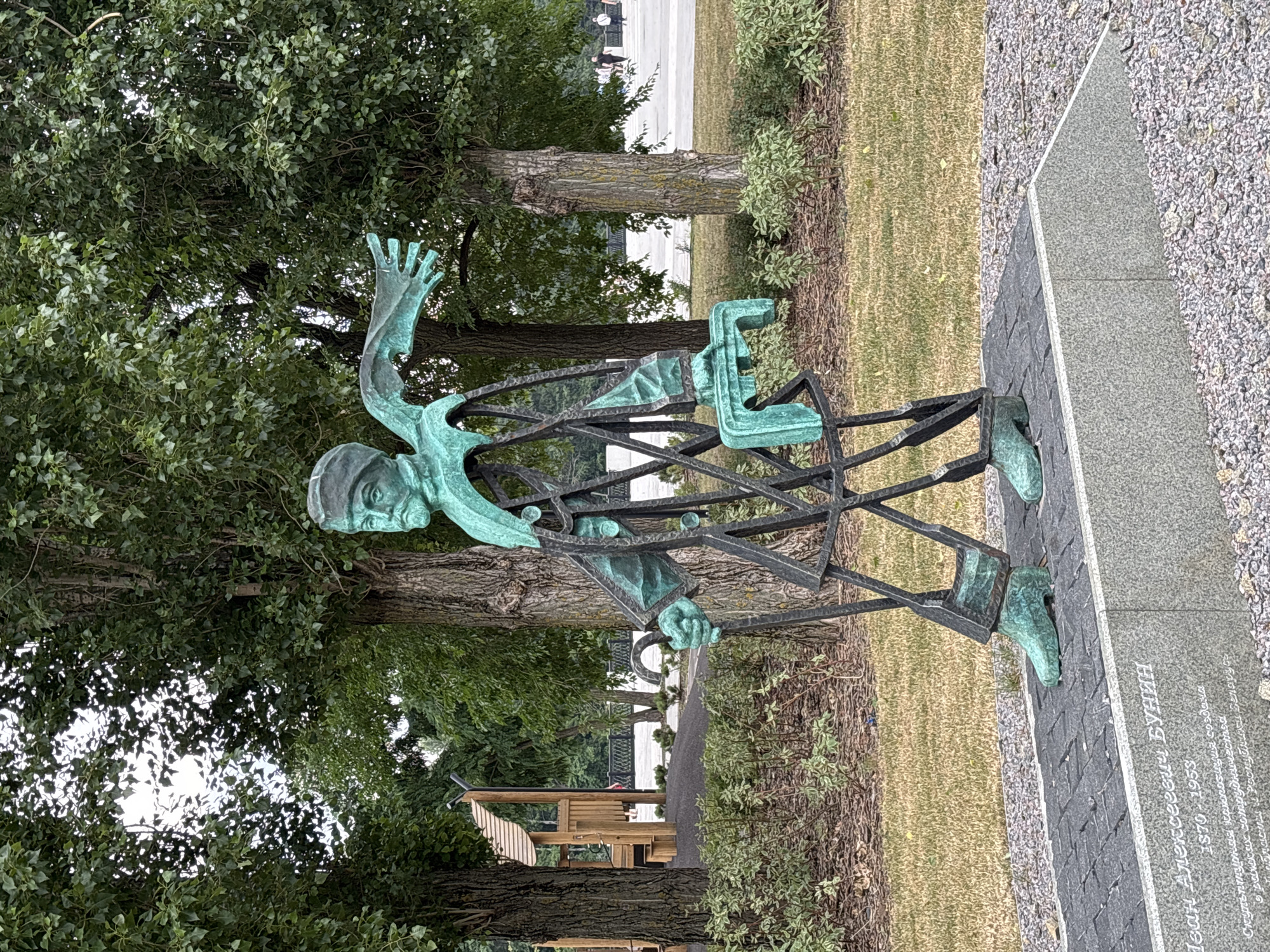 The image depicts a full-body, abstract bronze sculpture of a man in motion, situated outdoors in Novaya Usman', Russia. The sculpture is green-patinated and features a skeletal frame with stylized elements representing clothing and wings. The man appears to be walking, holding a cane in his left hand and carrying an object in his right. He wears a cap and has a beard. The sculpture is mounted on a dark gray, cobblestone-like base, which in turn rests on a larger, lighter gray granite pedestal. Inscribed on the pedestal, in white text, is "Иван Алексеевич Бунин 1870-1953" along with other text in Russian.
The sculpture is positioned in a park-like setting with a grassy lawn in the foreground and middle ground. Behind the sculpture, several large trees with full green foliage dominate the background. To the left of the sculpture, partially visible, is a wooden children's playground structure. In the far background, a paved area with several people in casual attire can be seen, and beyond that, a low fence and more trees. The sky is overcast, contributing to the soft lighting of the scene. The overall impression is of an artistic memorial in a public outdoor space.