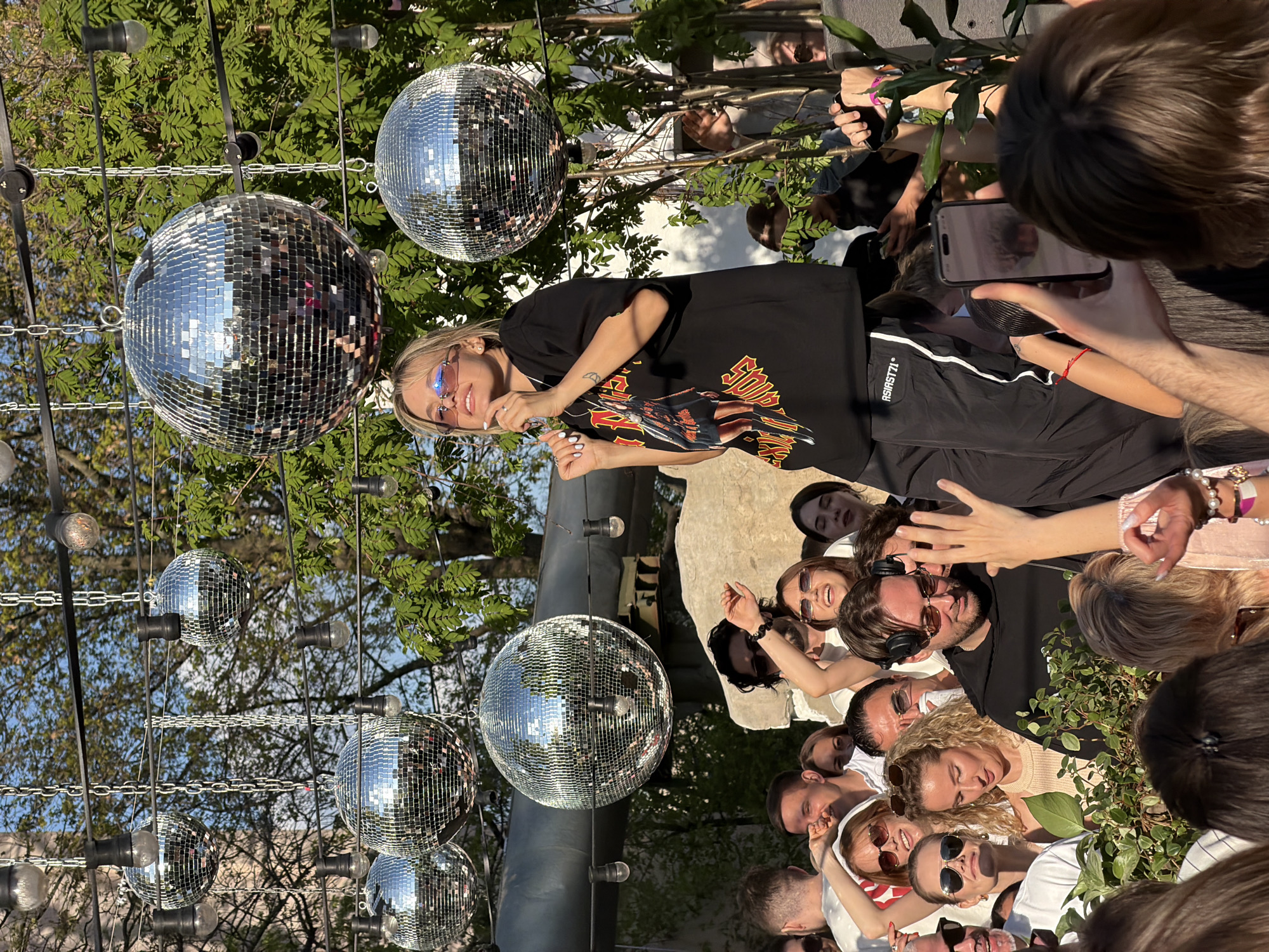The image shows a large group of people gathered outdoors in Moscow, Russia. Several people are clustered around a woman with blonde hair and sunglasses who is standing and speaking. Many of the people in the crowd are looking at her and appear to be listening. Some individuals are taking photos or videos with their phones.
Above the crowd hang multiple disco balls, suspended from a structure with lights. Trees and foliage are visible in the background, suggesting a park or garden setting. The general atmosphere appears celebratory or event-related.