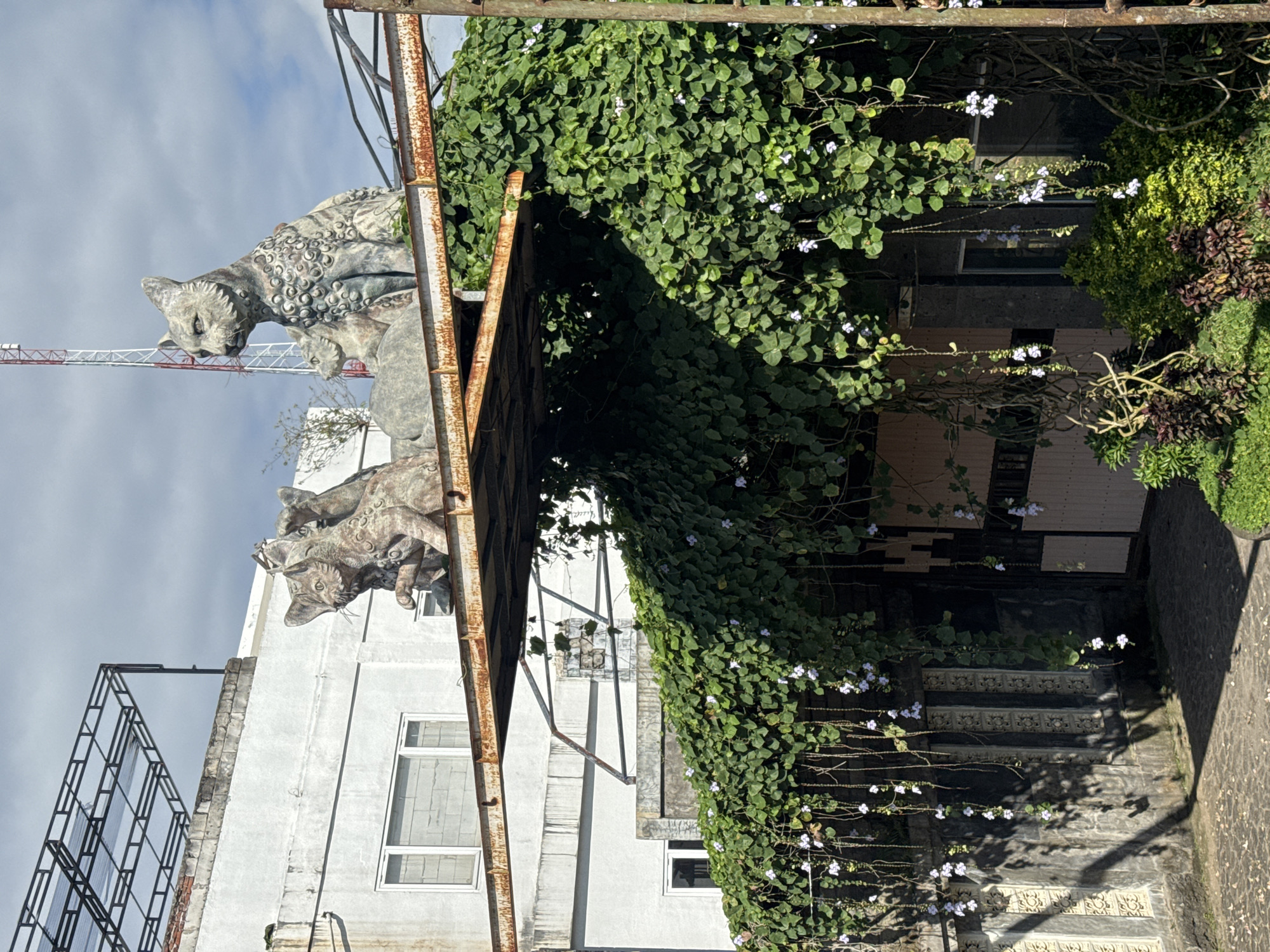 The image depicts an exterior view of a building in Kuta Selatan, Indonesia, identifiable by its architectural features and lush tropical vegetation. Atop the building's corner are decorative cat statues. The building's facade is partially covered with vines, featuring small white flowers. A rusty metal structure, possibly scaffolding or an awning frame, extends from the building. The building itself is white with visible windows. Part of an adjacent structure, likely a staircase, is visible with patterned detailing. The sky is partly cloudy. A red and white construction crane is visible in the background.