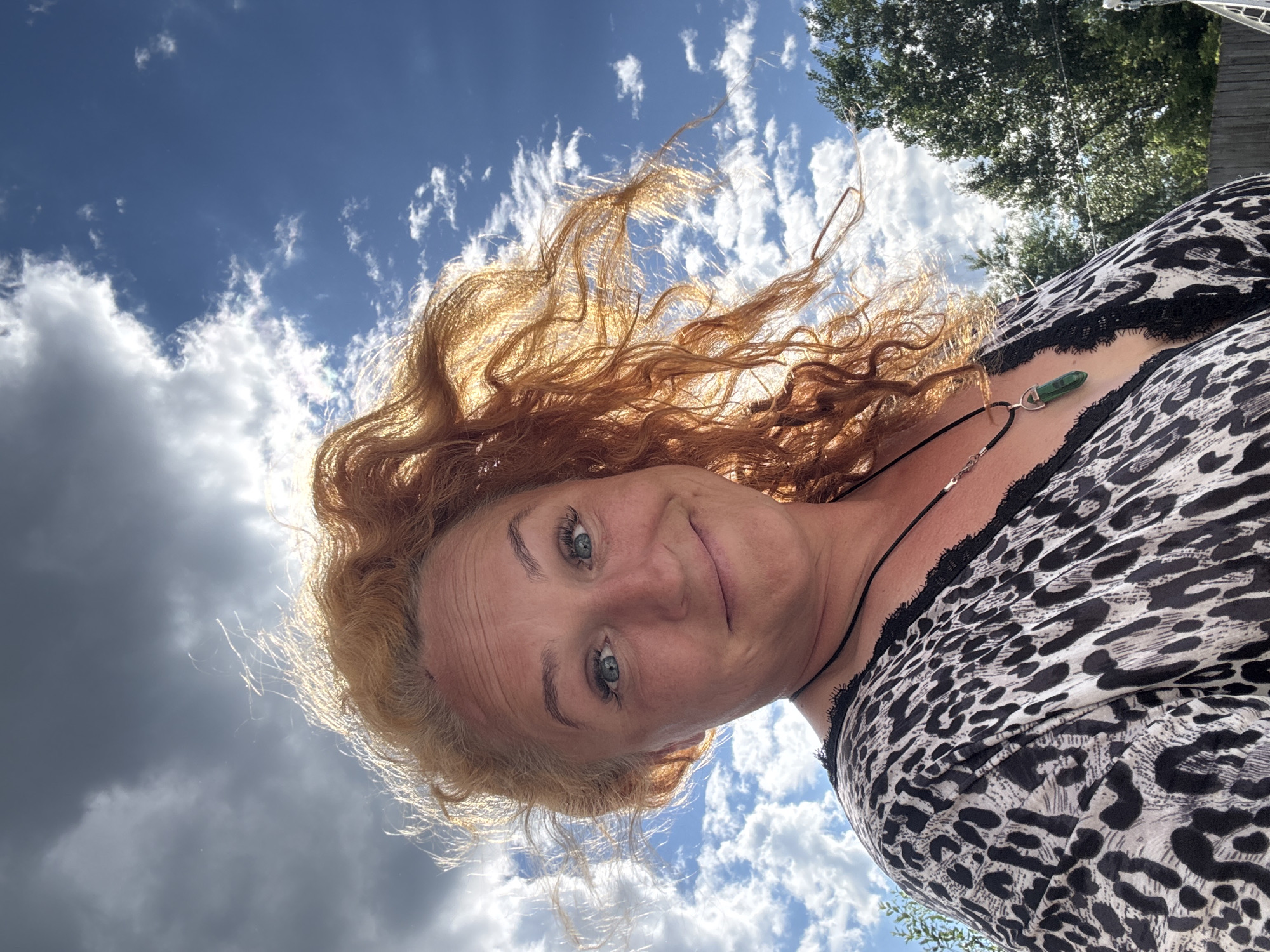 A woman with long, curly red hair is pictured from the chest up, taken from a low angle looking up towards a bright blue sky filled with white and dark clouds. Her hair is blowing in the wind, with some strands backlit by the sun. She is looking directly at the camera and smiling, with blue eyes and visible freckles. She is wearing a leopard-print top with black lace trim along the neckline and a black necklace with a green pendant. The background consists of the sky, some trees to the right, and a wooden fence. The image does not contain any information that would allow for its location to be determined, let alone specific to the city of Chaikovsky, Russia.