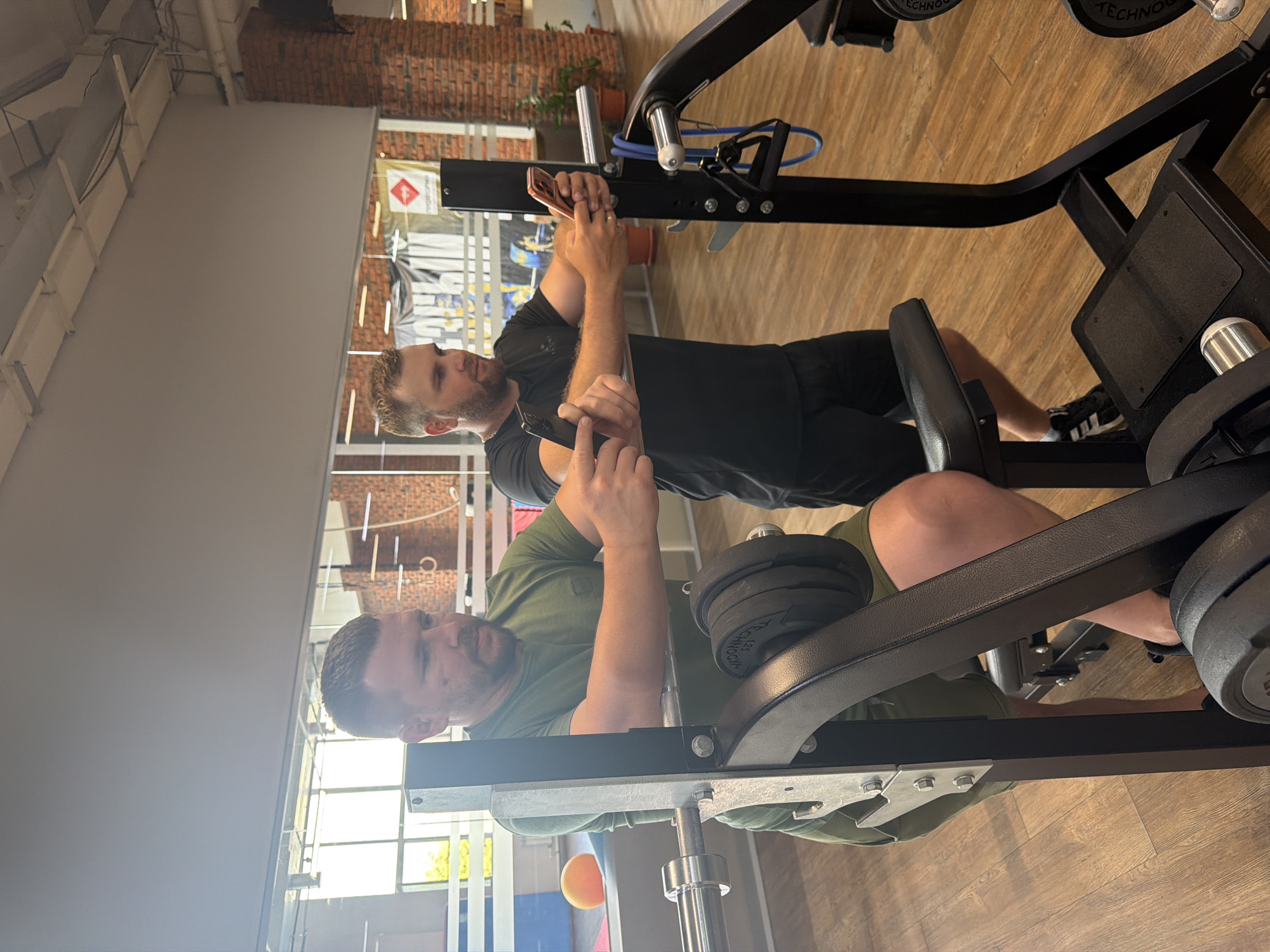 Two men are inside a gym, possibly located in Chaikovsky, Russia, as indicated by the context. Both men are holding smartphones and appear to be using them. The man on the left, wearing a green t-shirt and green shorts, is positioned near a weightlifting machine with weight plates attached. He is looking down at his phone, which he is holding in his right hand. The man on the right, wearing a black t-shirt and black shorts, is also looking at his smartphone, which he is holding in his right hand. He is standing next to the same weightlifting machine. The gym features wooden flooring and large windows. Brick walls are visible in the background, along with gym equipment and some decorative elements.