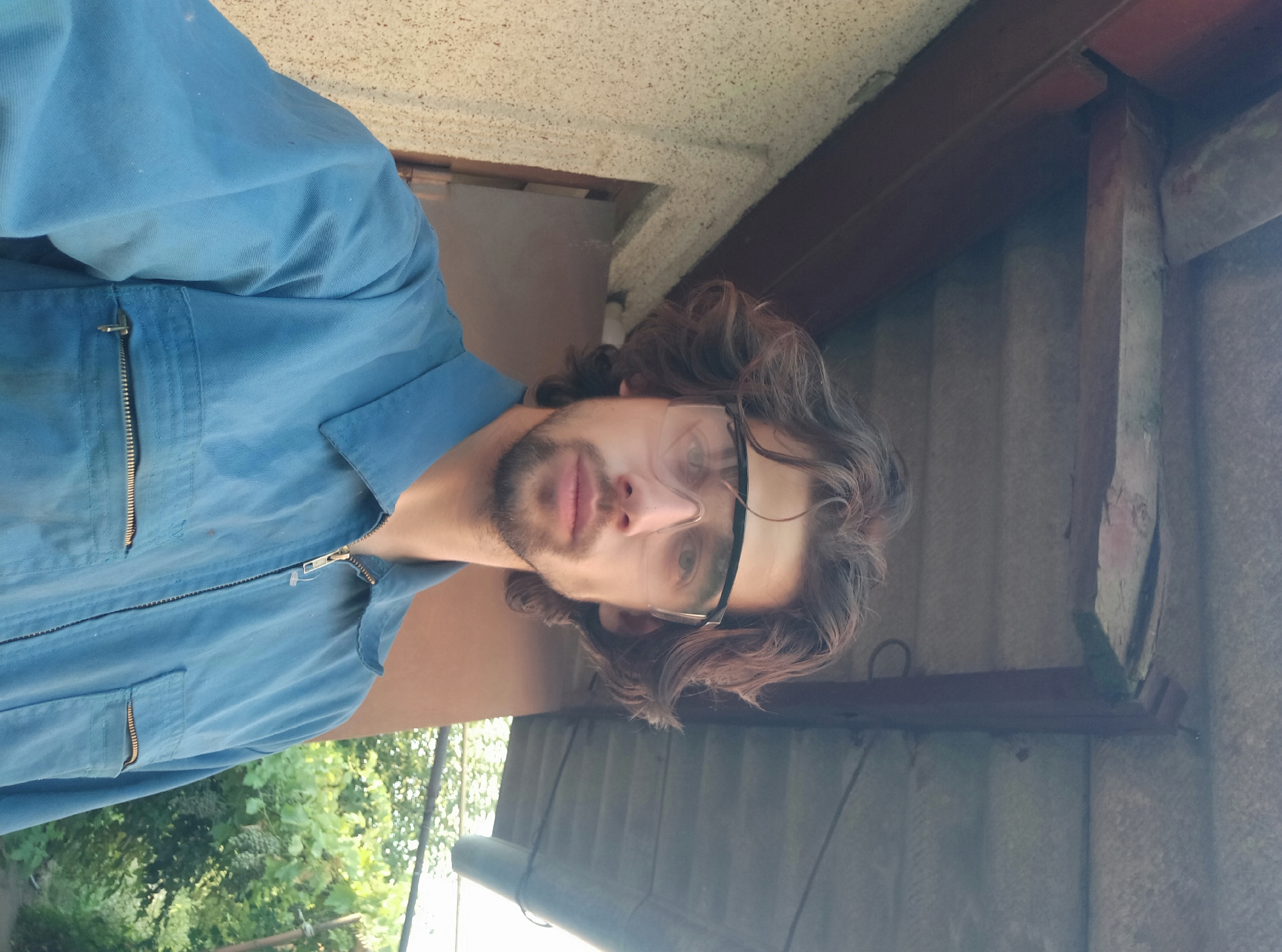 A man wearing blue coveralls and clear safety glasses is taking a selfie outdoors. He has medium-length, wavy brown hair and a short beard and mustache. The background includes a textured wall to the left, a brown door, and an overhanging roof structure made of wood and corrugated material. Lush green foliage and vines with grapes are visible behind him to the right. The setting appears to be a backyard or an outdoor workspace, potentially in Bender, Moldova, given the architectural style and vegetation.