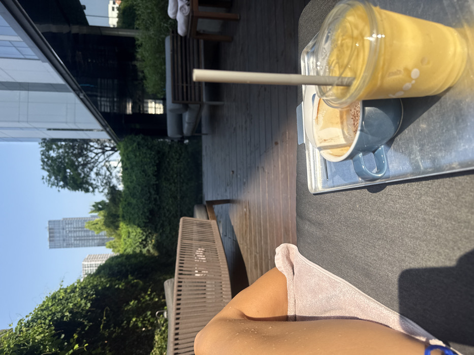 The image presents a first-person perspective of an outdoor lounge area in Bangkok, Thailand. In the foreground, the bare, tan legs of an individual are visible, exhibiting water droplets on the skin, resting on a dark grey cushioned surface partially covered by a light pink towel. A blue bracelet is visible on the left wrist. A metallic serving tray sits adjacent to the legs, holding a partially consumed light blue mug containing a coffee-like beverage and a clear plastic cup with a lid and a grey straw, filled with a yellow smoothie-like drink.
The midground features a dark wooden deck flooring. Two types of outdoor seating are present: a light brown woven lounge chair with a dark cushion to the left, and a dark grey slatted sofa-style lounge seat to the right. A small wooden side table with multiple neatly folded white towels is situated near the right lounge seat.
The background displays dense green foliage, likely hedges, separating the lounge area from the urban environment. Above the foliage, under a clear blue sky, several modern high-rise buildings are visible, including a prominent tall, slender skyscraper. A section of a contemporary building with glass panels and an angled roofline is also visible on the far right. Sunlight illuminates the scene, casting discernible shadows.