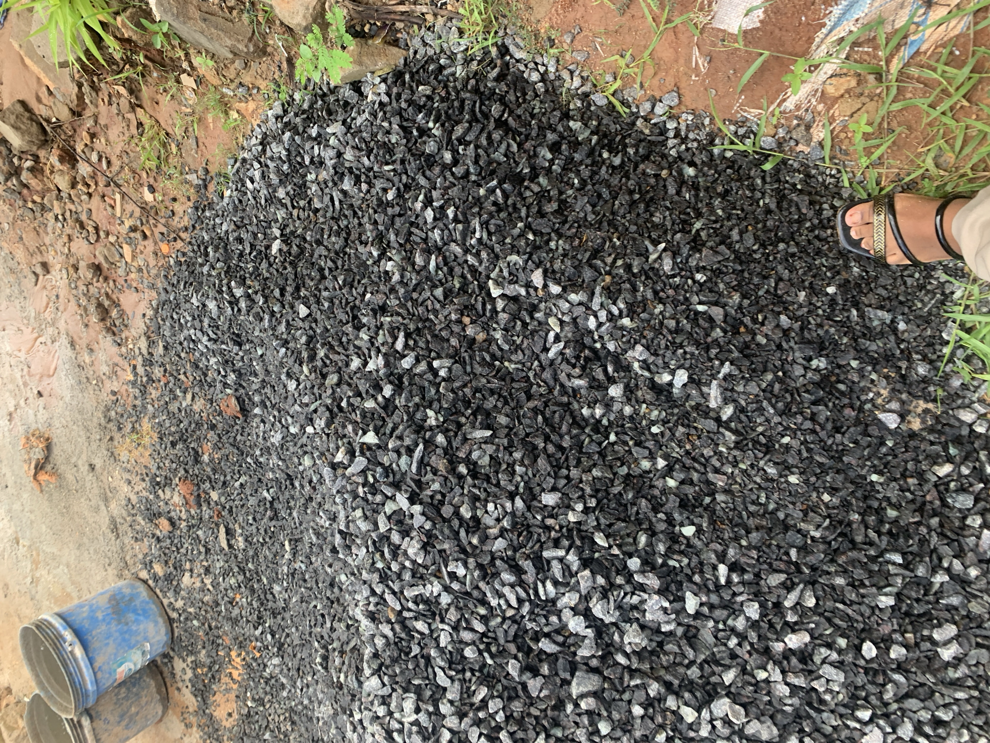 The image displays an outdoor ground-level scene in Dar es-Salaam, Tanzania, centered on a substantial pile of dark grey crushed aggregate. This aggregate occupies the majority of the frame's central and left portions. In the lower-right foreground, a bare human right foot, with dark skin, is visible, wearing a black sandal featuring a wide, textured gold-colored strap over the forefoot and a black ankle strap. A segment of light-colored legwear is also present above the ankle. The ground surrounding the aggregate pile is reddish-brown soil, marked by scattered patches of green grass and small plants, particularly on the right side. Several larger, natural rocks and fragments of a torn, woven material (possibly a sack) are embedded in or resting on this soil. In the upper-left background, two used and soiled plastic buckets are positioned, with the front bucket being blue and upright, resting on a lighter-colored, possibly concrete or compacted dirt surface. The overall scene suggests a construction or material storage area.
