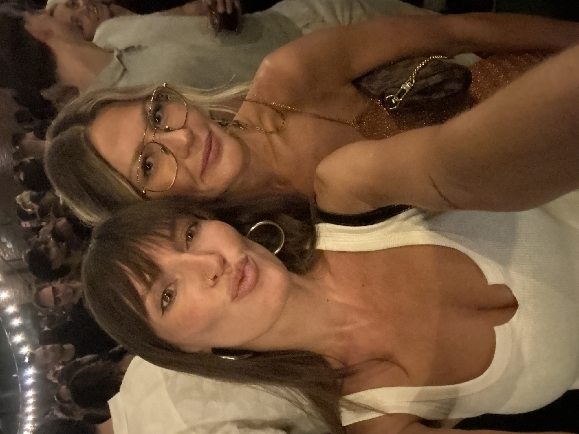 This selfie-style photo, captured in North Kuta, Indonesia, features two women in a dimly lit indoor setting, likely a social event or club, during the evening. In the foreground, a woman with dark brown hair and bangs is shown, looking directly at the camera with a playful pout. She wears large silver hoop earrings, a small nose piercing, and a white sleeveless top with a deep V-neck. Behind her, partially visible, is a blonde woman wearing gold-framed glasses and a subtle smile. She has a gold chain necklace and appears to be wearing a shimmery, possibly sequined, top. A small, dark handbag with a gold chain strap rests on her shoulder. The background is blurred, showing silhouettes of other people and small, bright lights, indicating a crowded and lively atmosphere. Both women appear relaxed and in good spirits. No text is visible in the image.