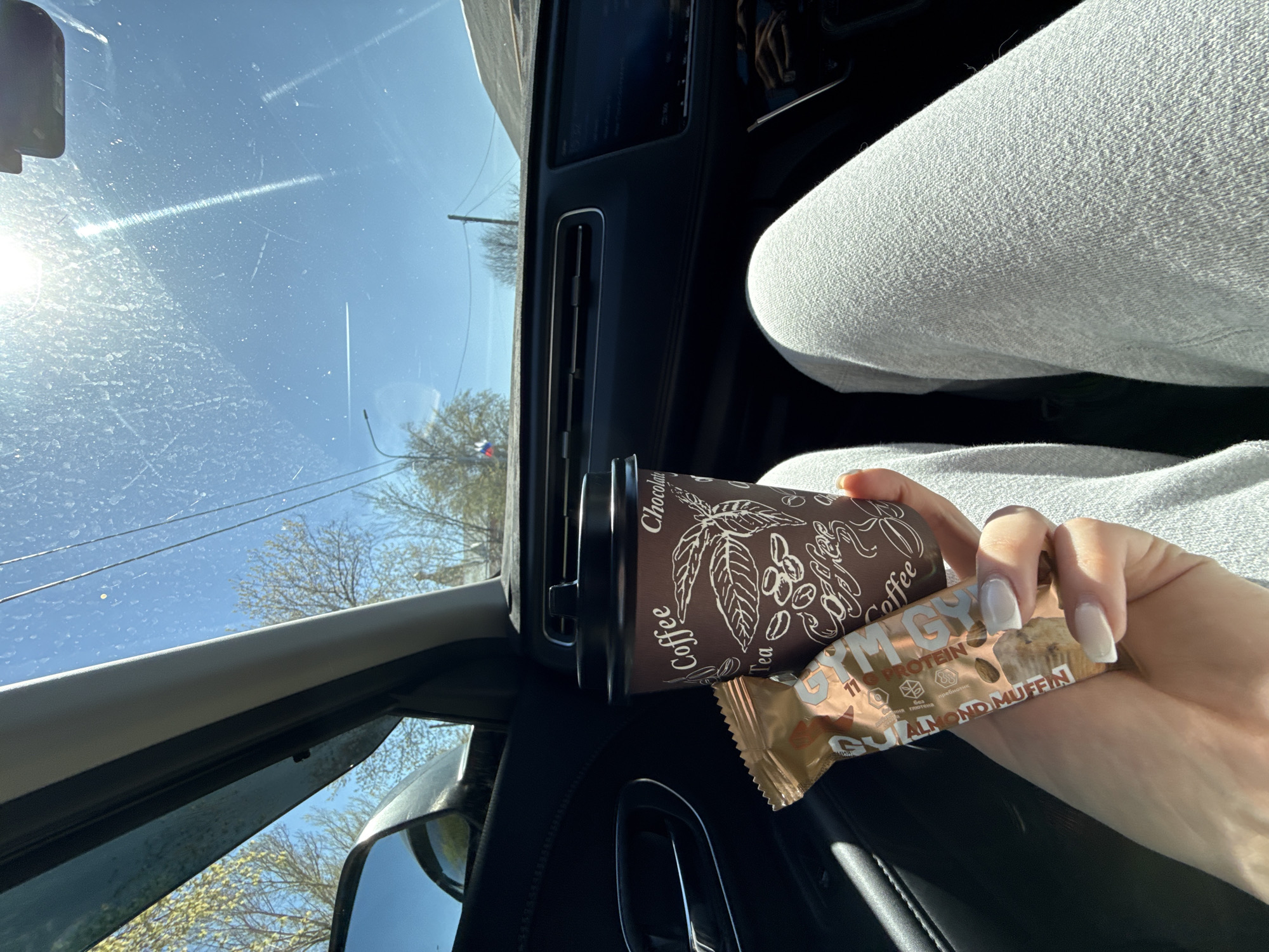 This media file captures a first-person perspective from inside a car on a clear, sunny day. The bright blue sky is visible through the windshield, which has some dust or streaks, with sun glare originating from the left. Outside the car, trees with sparse foliage and power lines suggest either late winter or early spring.
In the foreground, a person's hand, adorned with light-colored, square-tipped nails, holds a brown disposable coffee cup and a protein bar. The coffee cup is decorated with illustrations of coffee beans and leaves, featuring text such as "Coffee," "Chocolate," and "Tea." Next to it, the protein bar is wrapped in gold foil and branded "GYM GY." It prominently states "11g PROTEIN" and identifies the flavor as "ALMOND MUFFIN." Additional text in Russian, including "БЕЗ" (meaning "without"), is also visible on the wrapper.
The car's interior is partially visible, including a dark dashboard, an air vent, and a portion of a car seat. The person's leg, covered in light grey, textured fabric (likely sweatpants or casual trousers), is also seen. A rearview mirror is partially in view on the right side of the frame. The scene suggests a person consuming a quick snack and drink while on the go or taking a break in their vehicle.
