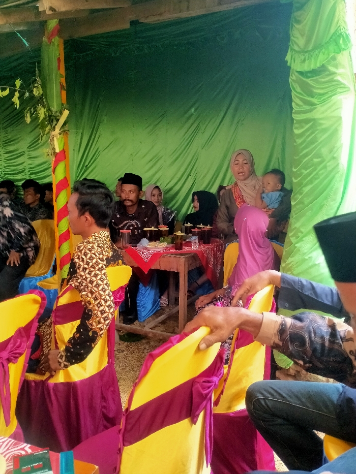 A gathering of people is seated around a wooden table covered with a red and white patterned cloth. On the table are small glass containers, likely filled with food or drink. The attendees include women in hijabs, a man in a dark hat, and a woman holding a baby. Yellow chairs with burgundy accents line the foreground, while a green fabric backdrop creates a festive atmosphere. The scene appears to be an informal celebration or event.