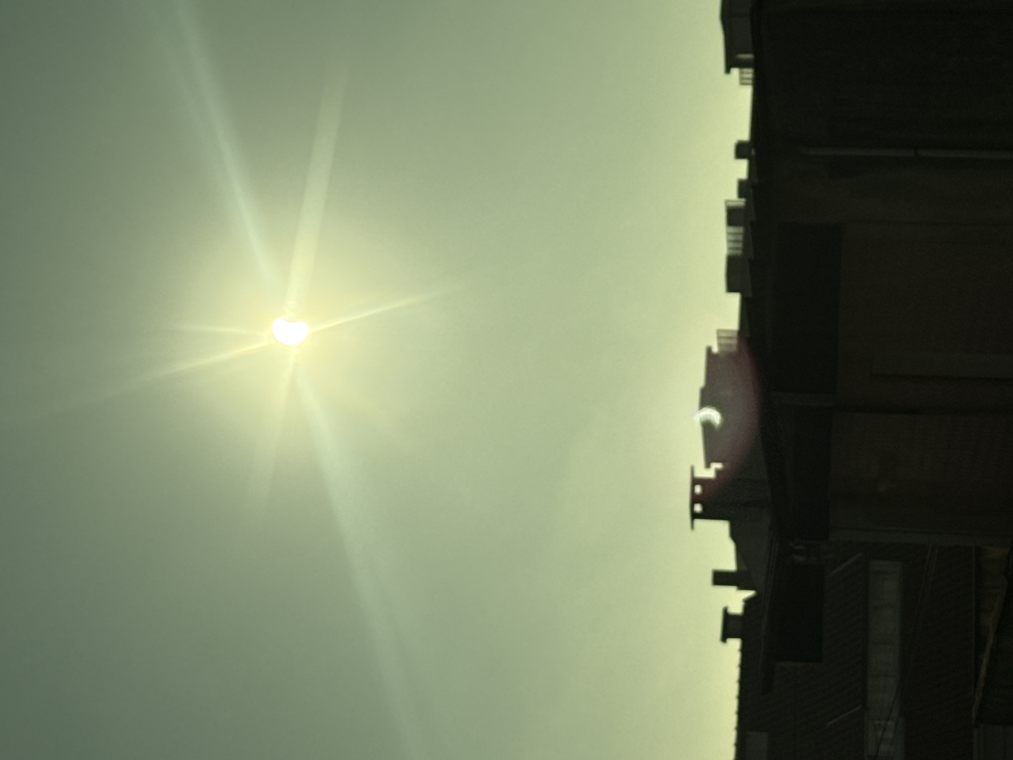 This media file captures a partial solar eclipse over Vila Nova de Gaia, Portugal. The sun, partially obscured by the moon, emits bright rays that create a halo effect against a pale green sky. The celestial event is observed from the perspective of a building. The right side of the image is dominated by the dark silhouette of a multi-story structure, possibly an apartment building or industrial facility, with visible balconies and architectural details. The building's dark facade contrasts sharply with the bright sky. The time of day appears to be late morning or early afternoon, given the position of the sun. The weather seems clear, as the sun is visible despite the eclipse. The scene evokes a sense of wonder and a rare astronomical occurrence. There is no visible text in the image.