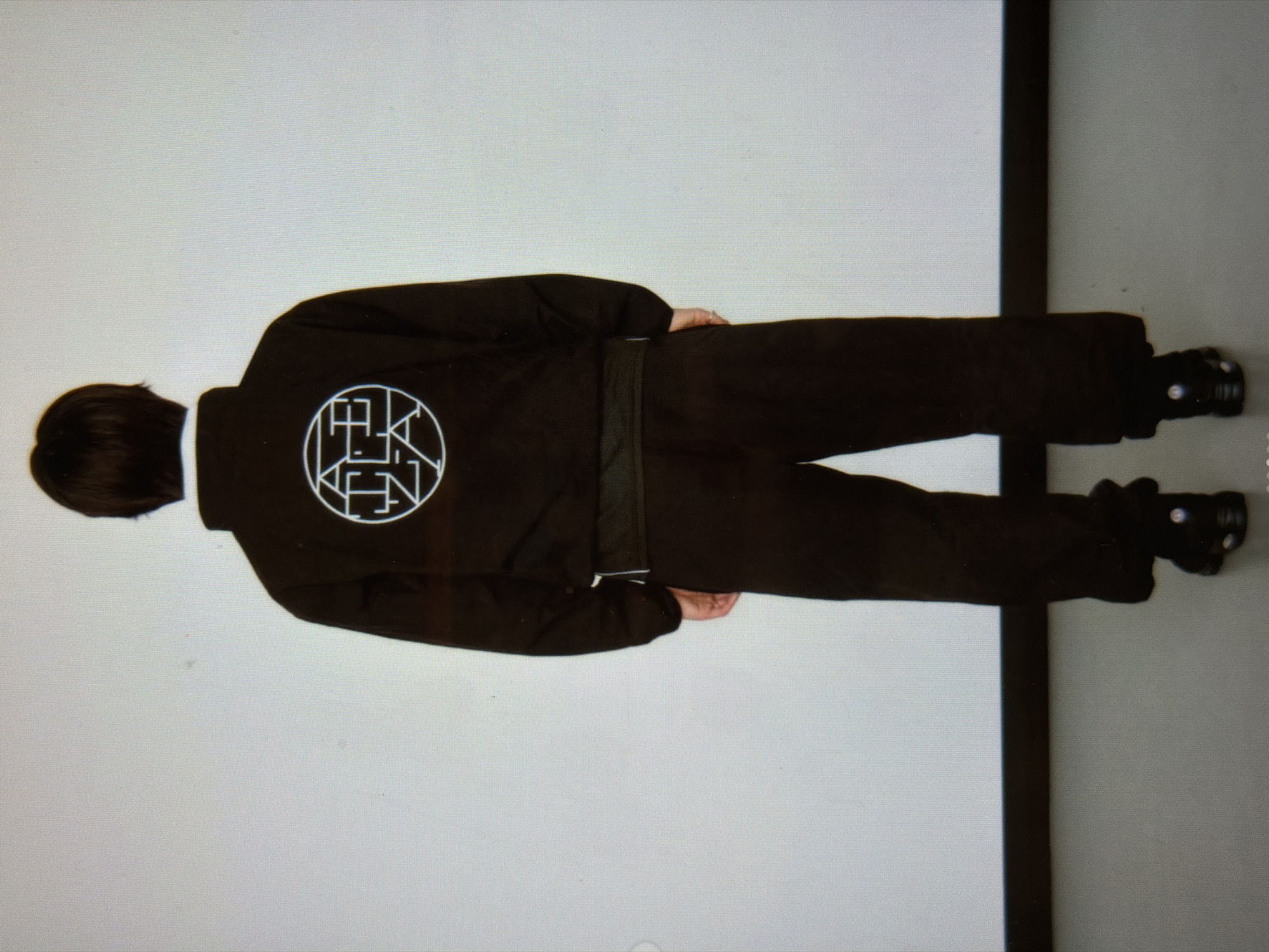 A person stands with their back to the viewer, wearing a dark, long-sleeved jacket and pants that appear to be a matching set. A white circle with stylized lettering is visible on the upper back of the jacket. The person is wearing dark shoes. They are positioned against a wall with a visible color gradient; the left side of the wall is lighter than the right side. The setting appears to be indoors. There is no information available in the image to suggest the location is New York City or any specific city.