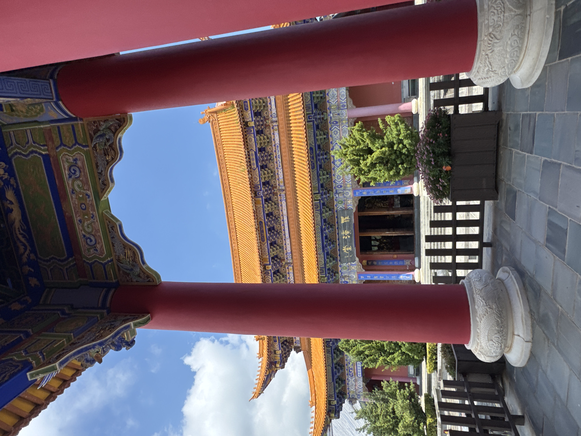 The image depicts a view through a traditional Chinese temple's red pillars towards its ornate facade and tiered, orange-tiled roof. The temple architecture features intricate carvings and colorful painted details. In front of the temple, there are green trees and a planter box filled with pink flowers. The ground is paved with dark gray stone tiles. The sky is blue with scattered white clouds. The scene is calm and appears to be outdoors during the daytime. The temple is likely part of the Three Pagodas of Chongsheng Temple in Dali City, China, a UNESCO World Heritage site.