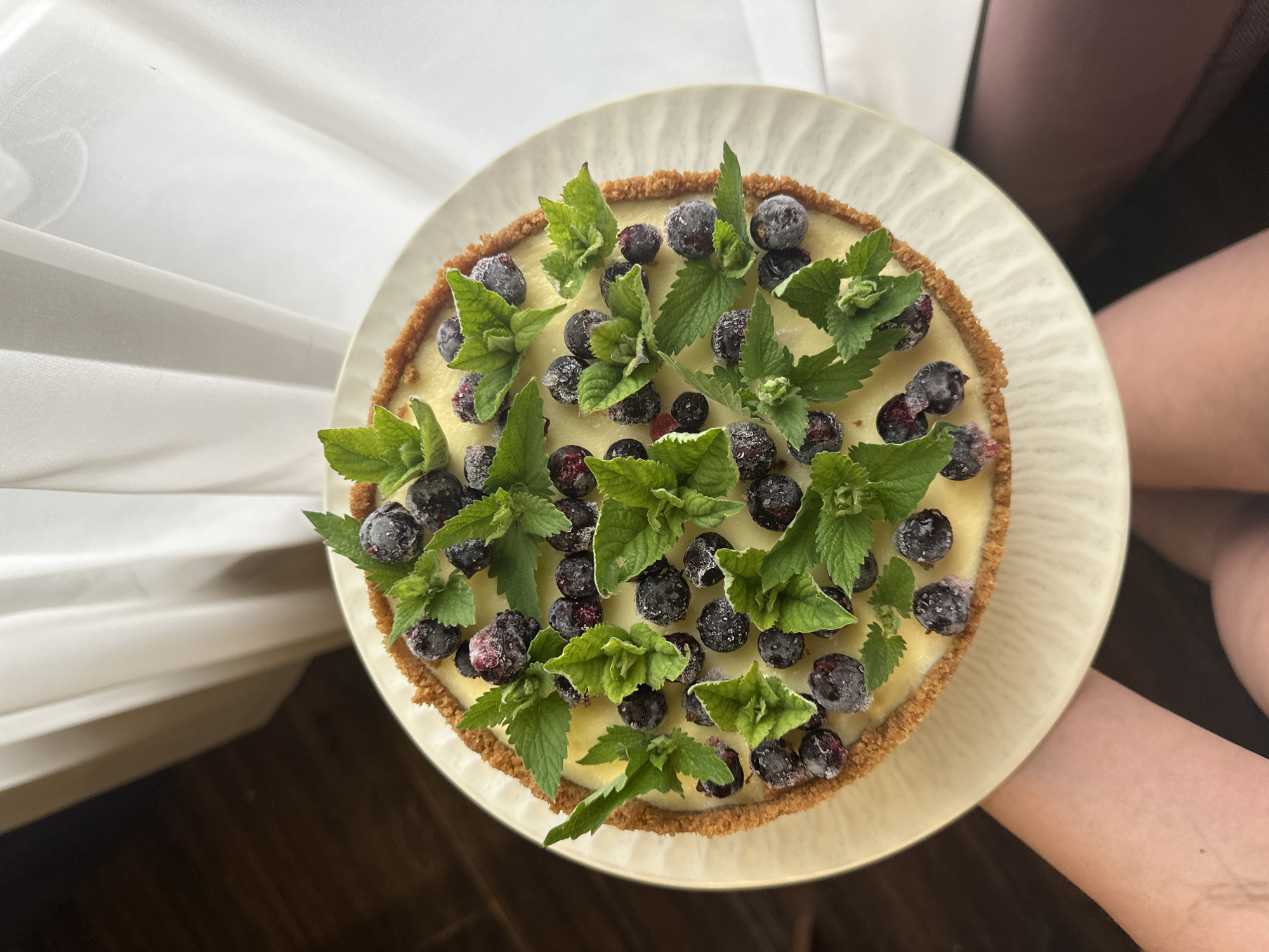 The image presents a top-down, slightly angled view of a round dessert, likely a tart or cheesecake, centered on a cream-colored, circular plate with a subtly ridged rim. The dessert features a smooth, pale yellow filling contained within a textured, light brown crumb crust that forms the perimeter. The top surface is generously garnished with numerous dark blue-purple berries, consistent with frozen blueberries or blackcurrants, which exhibit a frosty or sugar-dusted appearance. Interspersed among the berries are multiple clusters of vibrant green, fresh mint leaves. The plate is partially supported by light-skinned hands, visible at the lower left and right edges of the frame. The background to the upper left is an out-of-focus dark brown surface, while the right and upper right background consists of soft, white, sheer curtains, suggesting an indoor setting illuminated by natural light. No discernible visual elements within the image provide confirmation of its specific location in the city of Chaikovsky, Russia.