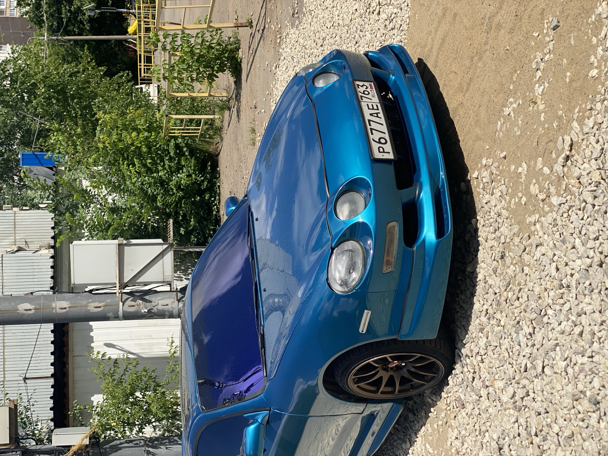 The image shows a bright blue, sporty car parked on a gravel surface. The car has a customized body kit and tinted windows. A license plate is visible on the front of the car with the text "P677AE 763 RUS". The scene is outdoors, with green trees and a building in the background. A yellow metal structure, possibly a playground or fence, is also present. The sun is shining, casting shadows, suggesting it is daytime. The gravelly ground and the presence of trees indicate an outdoor, possibly suburban or semi-rural, location. There are no people visible in the image. The overall impression is of a sunny day with a focus on the parked vehicle. The image appears to be taken in Kotelniki, Russia.