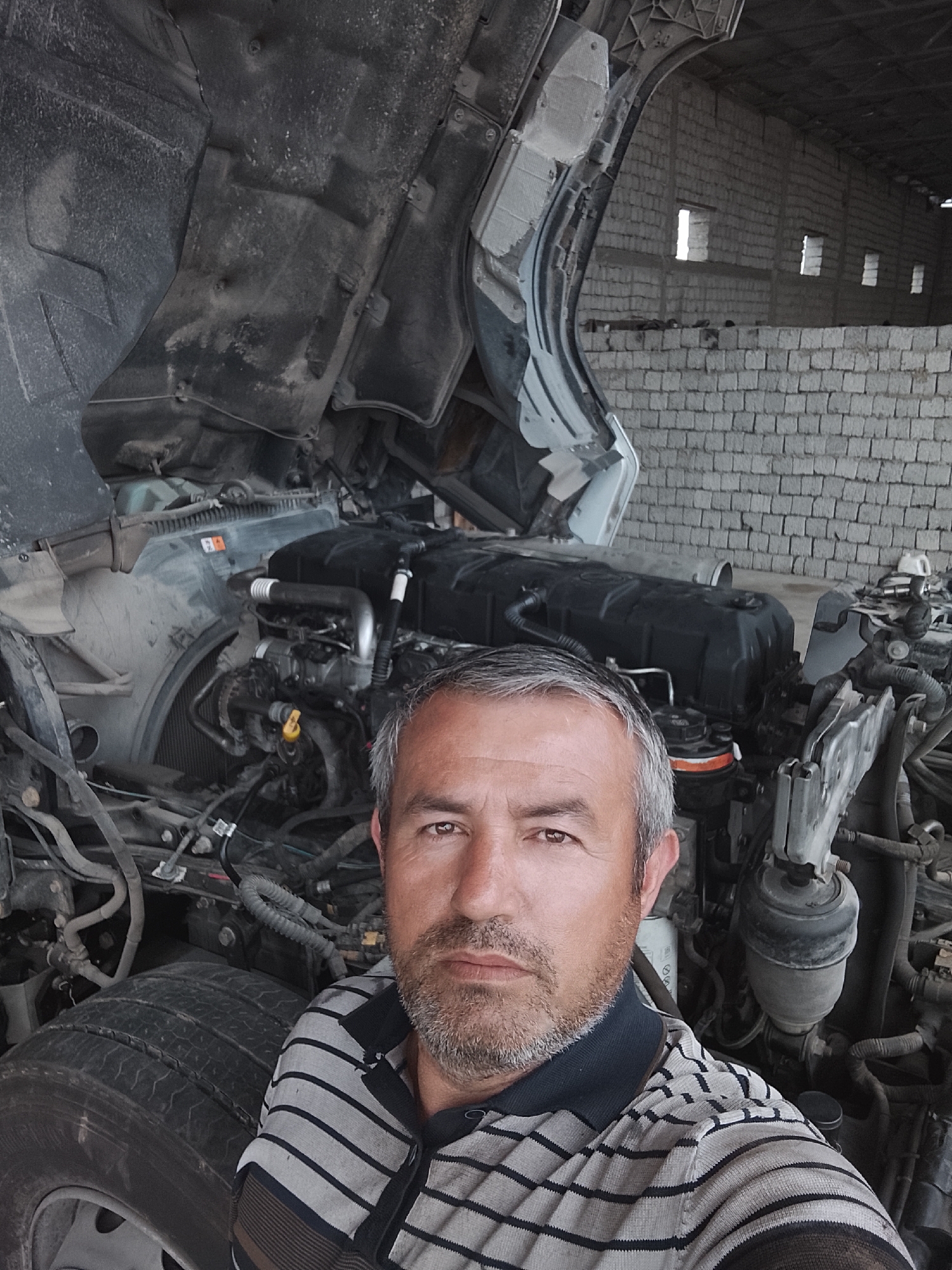 A middle-aged man stands in front of a large, open-hooded truck, presumably in a garage or maintenance facility. The man has short, grey hair and a salt-and-pepper beard, wearing a striped shirt. The truck's engine is exposed, showing various mechanical components. The background features a wall constructed of light-colored cinder blocks with small windows. The photograph was taken in Sabir Rakhimov, Uzbekistan.