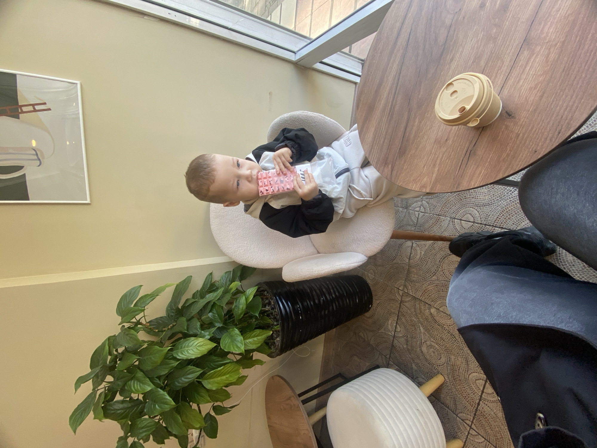 A young boy in a black and white tracksuit sits in a white, round chair, holding a pink and white patterned object. The chair is next to a round wooden table with a coffee cup on it. A potted green plant is nearby. The floor is tiled in a circular pattern. A window is in the background, and a modern art piece is on the wall. Another chair with someone sitting in it is adjacent to the table. It is impossible to determine that this photo was taken in Sochi, Russia.