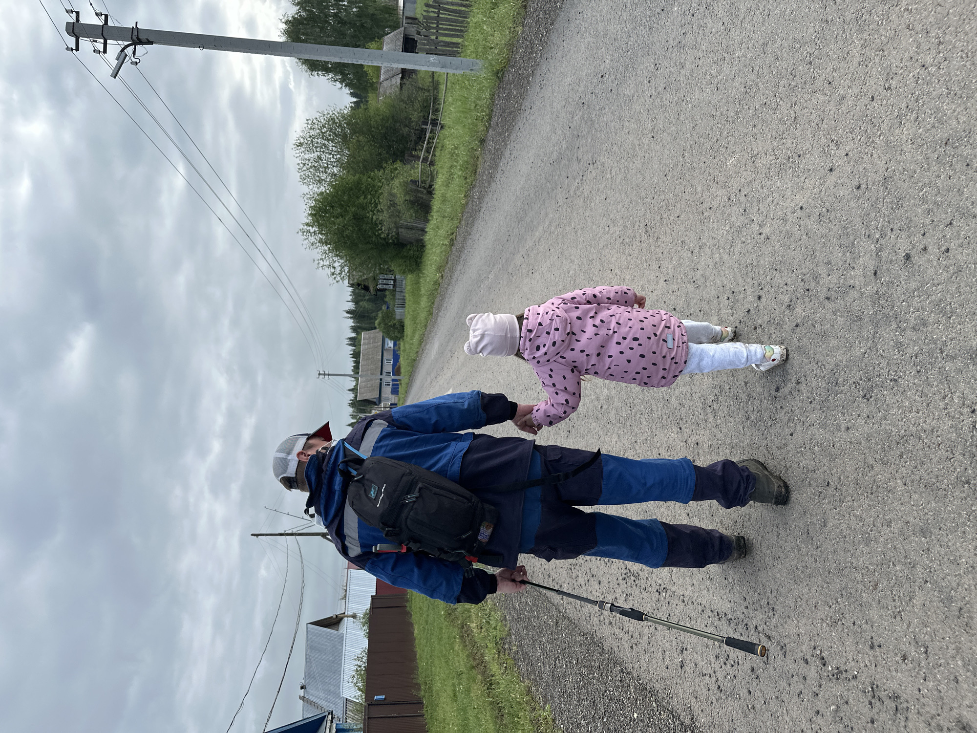 In the village of Среднее Кечево, Russia, a man and a young child walk hand-in-hand down a paved road. The man, seen from the back, wears a two-tone blue jacket and pants, a baseball cap, and a backpack. He holds a walking stick. The child, also seen from the back, wears a pink jacket with black polka dots, white pants, and a white hat with bear ears. Utility poles and wooden houses line the side of the road. The sky is overcast.