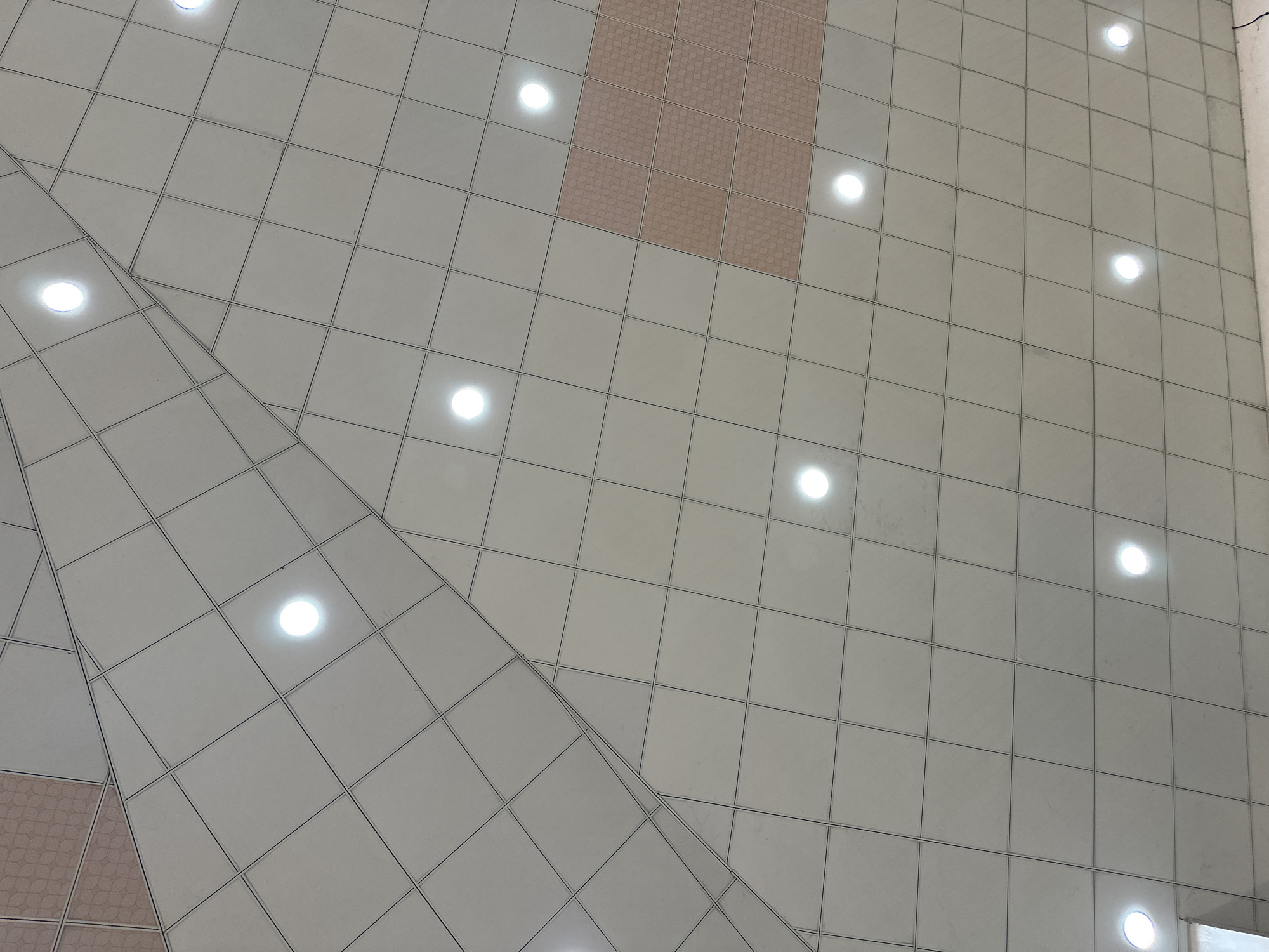 The image shows a section of a modern ceiling in Jalingo, Nigeria. The ceiling is composed of square panels, predominantly white or light gray, arranged in a grid pattern. These panels are intersected by darker lines, indicating the seams between them. Several circular lights are embedded within the panels, emitting a bright, white glow. In some areas, there are rectangular panels with a textured, light pink or beige pattern, featuring a repeating circular design. The ceiling appears to be part of a large indoor space, possibly an airport, shopping mall, or convention center, given its scale and contemporary design. The lighting suggests an artificial environment, characteristic of such public indoor venues. No people or specific actions are visible.