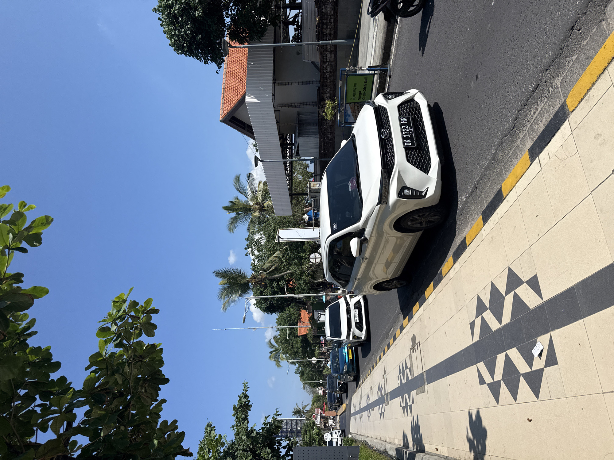 The image captures a bright, sunny street scene in what appears to be Kuta, Indonesia, given the context. The time of day is midday, indicated by the strong shadows and clear, bright blue sky.
In the foreground, a detailed sidewalk features a decorative pattern of alternating light and dark geometric triangles, bordering a curb painted with a yellow and black stripe. The street itself is asphalt.
A prominent white car, possibly an SUV or crossover, is visible in the midground, either parked or slowly moving on the street. Its Indonesian license plate, "DK 1723 HR" with "11-27" below, is clearly legible on the front. Behind this car, other vehicles, including another white car and a blue car, can be seen further down the road, suggesting a busy thoroughfare.
To the right of the white car, a sign with green and white text is visible, reading "KERANJANG BALI," "Surga," and "Oleh-Oleh Bali," indicating a local souvenir shop. Beyond the street, buildings with traditional reddish-brown tiled roofs and white facades are visible. The area is lush with tropical foliage, including tall palm trees and broadleaf trees, contributing to the bright, tropical setting. The weather is clear and sunny with no visible clouds. No specific activity or event is taking place, but rather a typical daytime street environment.