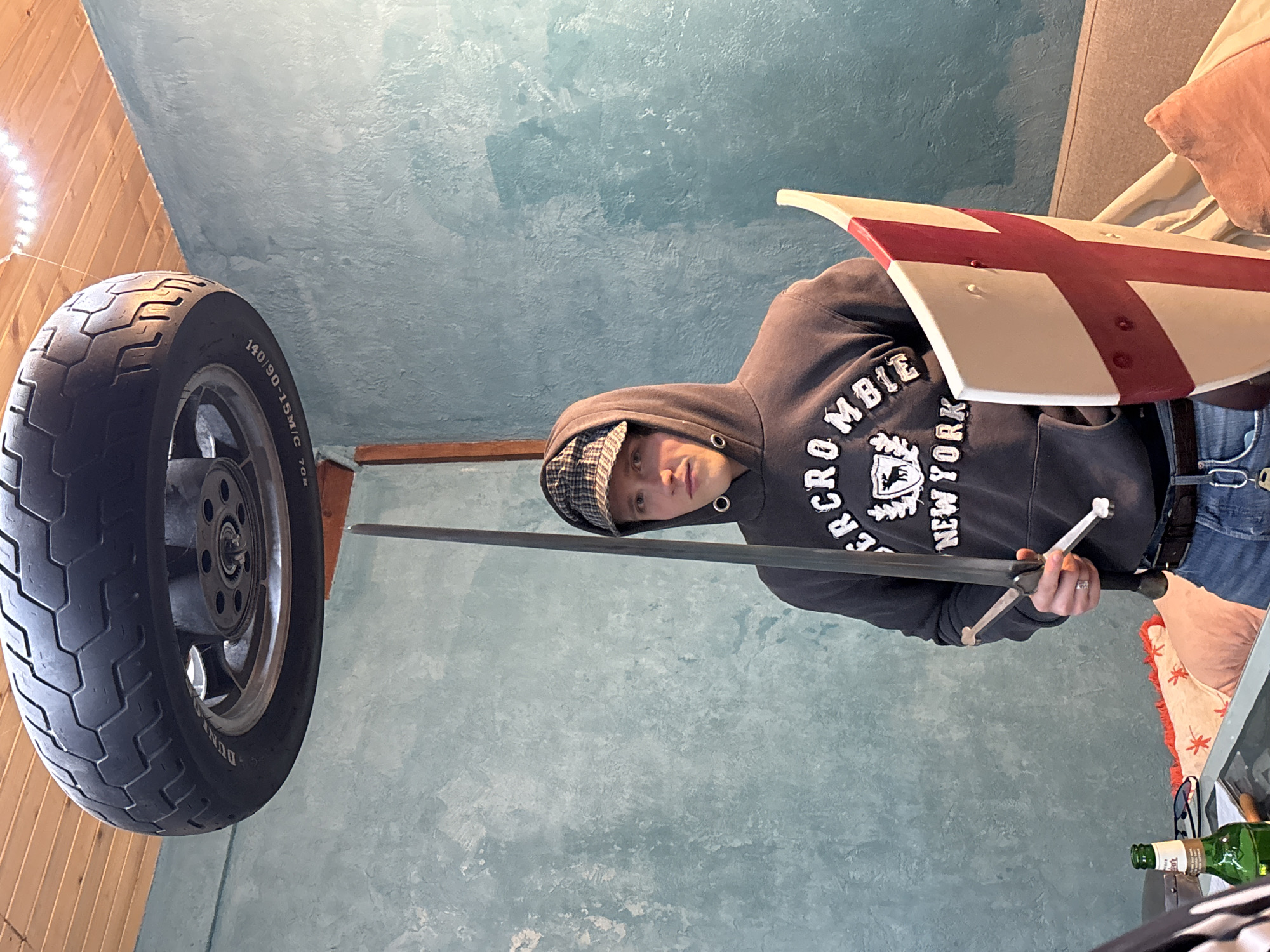 The photograph shows a person in a gray Abercrombie & Fitch hoodie and a checkered cap holding a sword and shield with a red cross design. The person is positioned in front of a textured blue wall, with a motorcycle tire and rim visible in the background. The environment includes wooden paneling on the left and a glimpse of a sofa with pillows on the right. A glass bottle is visible on a surface to the right. There is no evidence to suggest the location is in Verinsko, Bulgaria.