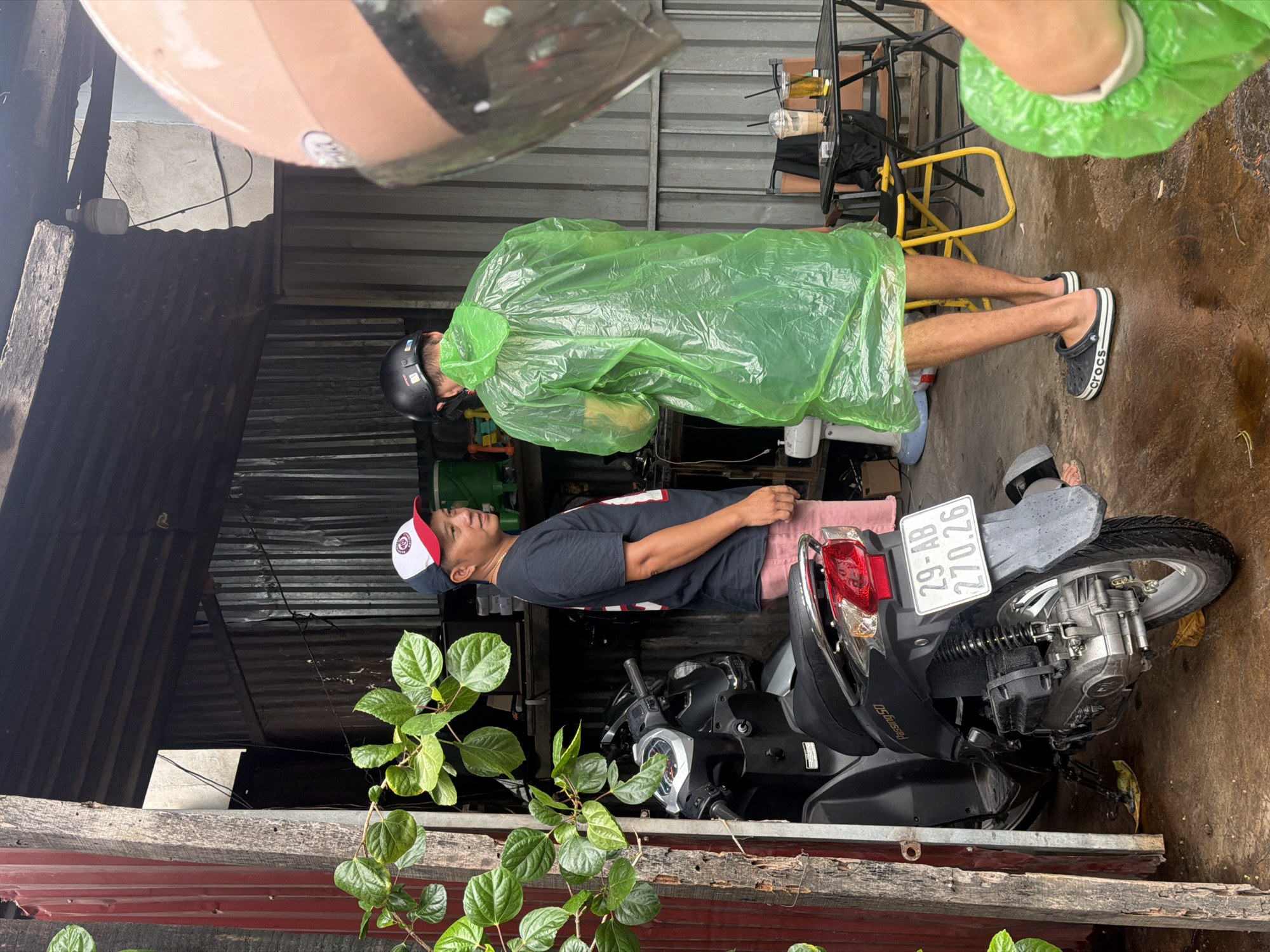 The image depicts a street scene in Nha Trang, Vietnam, likely in a residential or commercial alleyway. In the foreground, a black scooter with the license plate "29-AB 270.26" is parked. To the left, green foliage hangs from a wooden structure. Two men are engaged in conversation. One man, wearing a black t-shirt, red and white cap, and pink shorts, stands near the scooter. The other man, dressed in a bright green disposable raincoat and black helmet, faces away from the camera. In the background, a corrugated metal structure forms a covered area. A small table with two drinks and cigarettes is visible. The ground is wet, suggesting recent rain. The overall impression is a casual, everyday moment captured in a utilitarian urban environment.