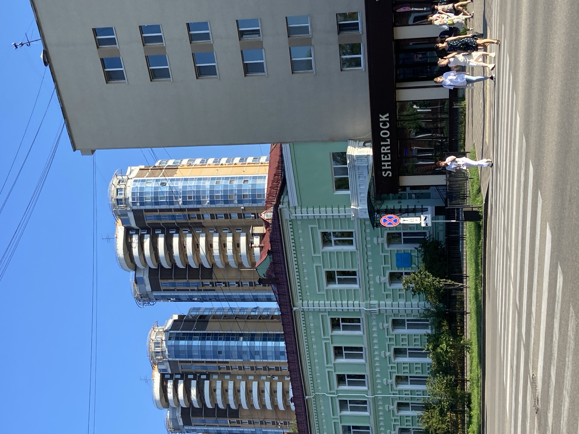 This image depicts a street scene in Blagoveshchensk, Russia, likely on a sunny day, given the clear blue sky and shadows. The foreground features a pedestrian crosswalk with several people crossing. To the right of the crosswalk is a modern building with the sign "SHERLOCK" visible above its entrance. Behind this building, and slightly to the left, is a lower, light green building with white trim and numerous windows. In the background, several tall, modern apartment buildings with curved balconies and glass facades dominate the skyline. Overhead, power lines are strung across the sky. The overall impression is of a city intersection where older and newer architectural styles coexist.