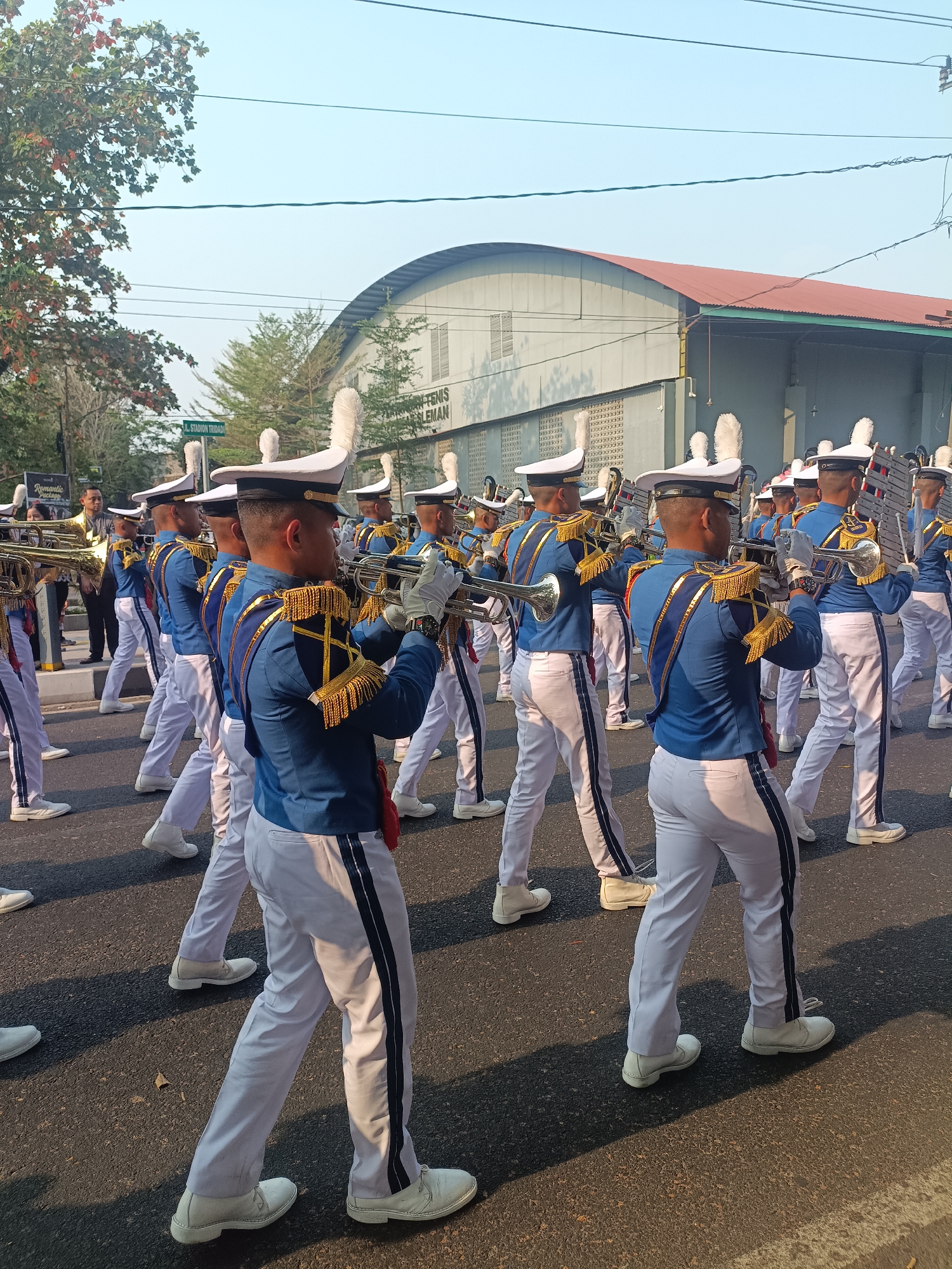 A marching band, comprised of young men wearing blue uniforms with gold trim and white hats, plays brass instruments on a paved street. The musicians are in formation, marching towards the right of the frame. In the background, a large, arched building with a red roof and grey walls is visible. Above the building, power lines crisscross against a clear, light blue sky. Trees with green and reddish foliage are on the left side of the image. A street sign visible in the distance reads "JL. STADION TRIDADI SLEMAN". The lighting suggests it is daytime, likely late afternoon given the long shadows cast on the road.