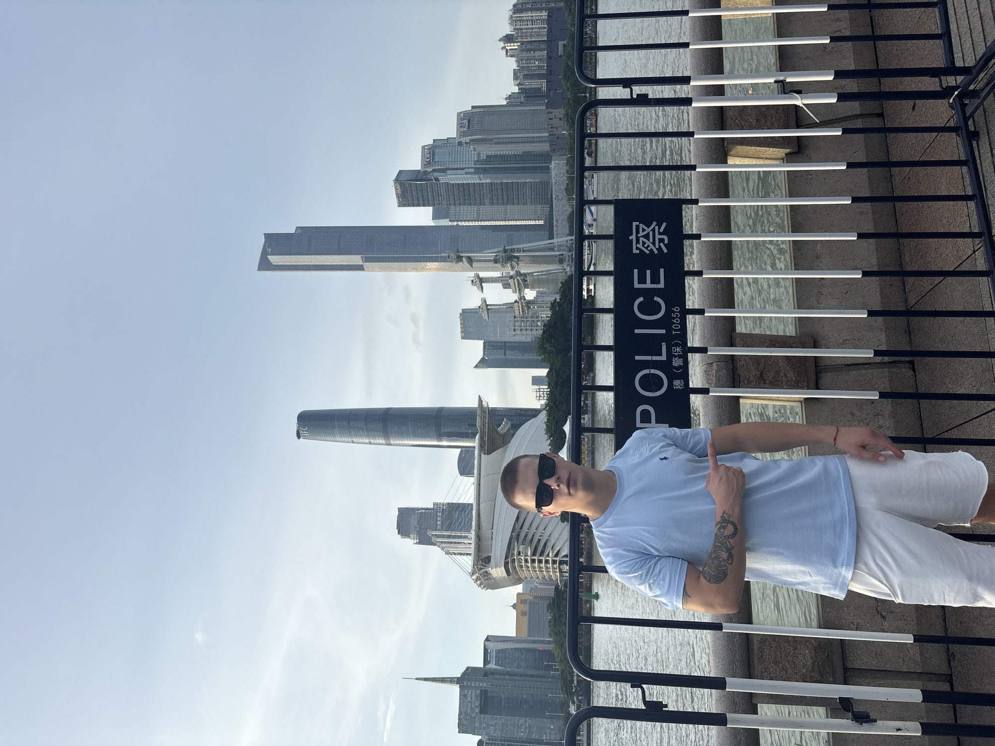A young Caucasian male stands in front of a body of water, pointing towards a black sign with white lettering that reads "POLICE". The man is wearing a light blue t-shirt, white shorts, and sunglasses. He has a tattoo of a tiger on his left forearm. Behind him, a cityscape of tall buildings, including the Guangzhou IFC and CTF Finance Centre, is visible. The scene appears to be along the Haizhu District waterfront in Guangzhou, China. The sky is overcast, and the water is calm. There are metal barriers in the foreground, separating the man from the water.