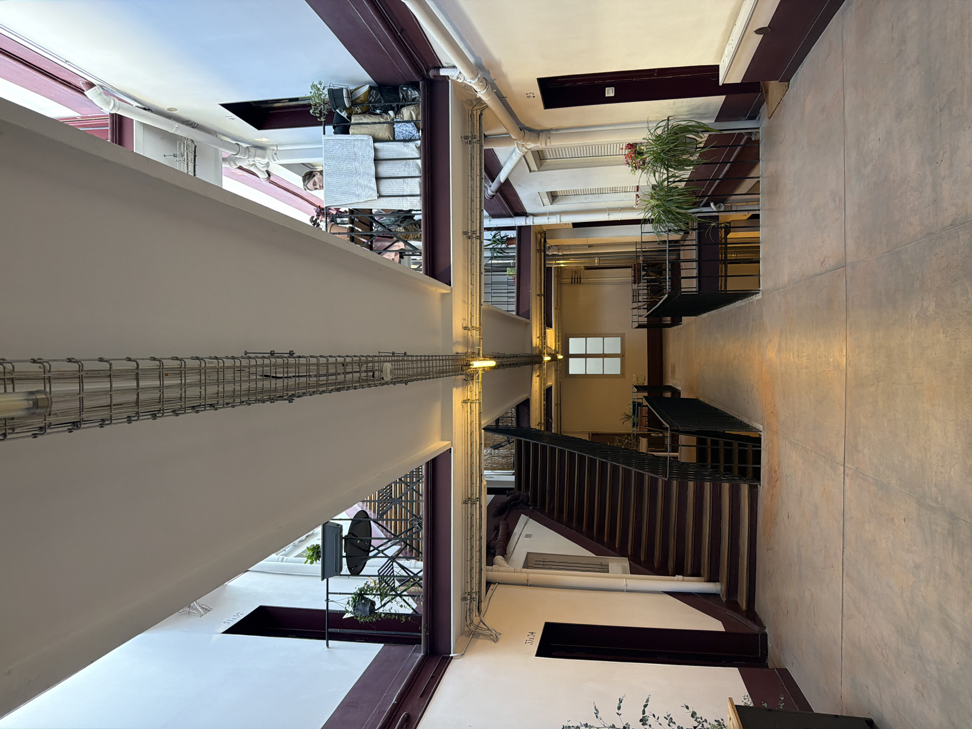 The image depicts an interior space in Paris, France, possibly a former industrial building that has been repurposed, as evidenced by the exposed piping and metal support structures. The architecture is angular with a mix of white walls, dark burgundy trim, and concrete floors. A central void runs the length of the space, connecting different levels. There are staircases with metal railings that lead between floors. Plants are visible on multiple levels and in various locations, adding a touch of greenery to the industrial setting.