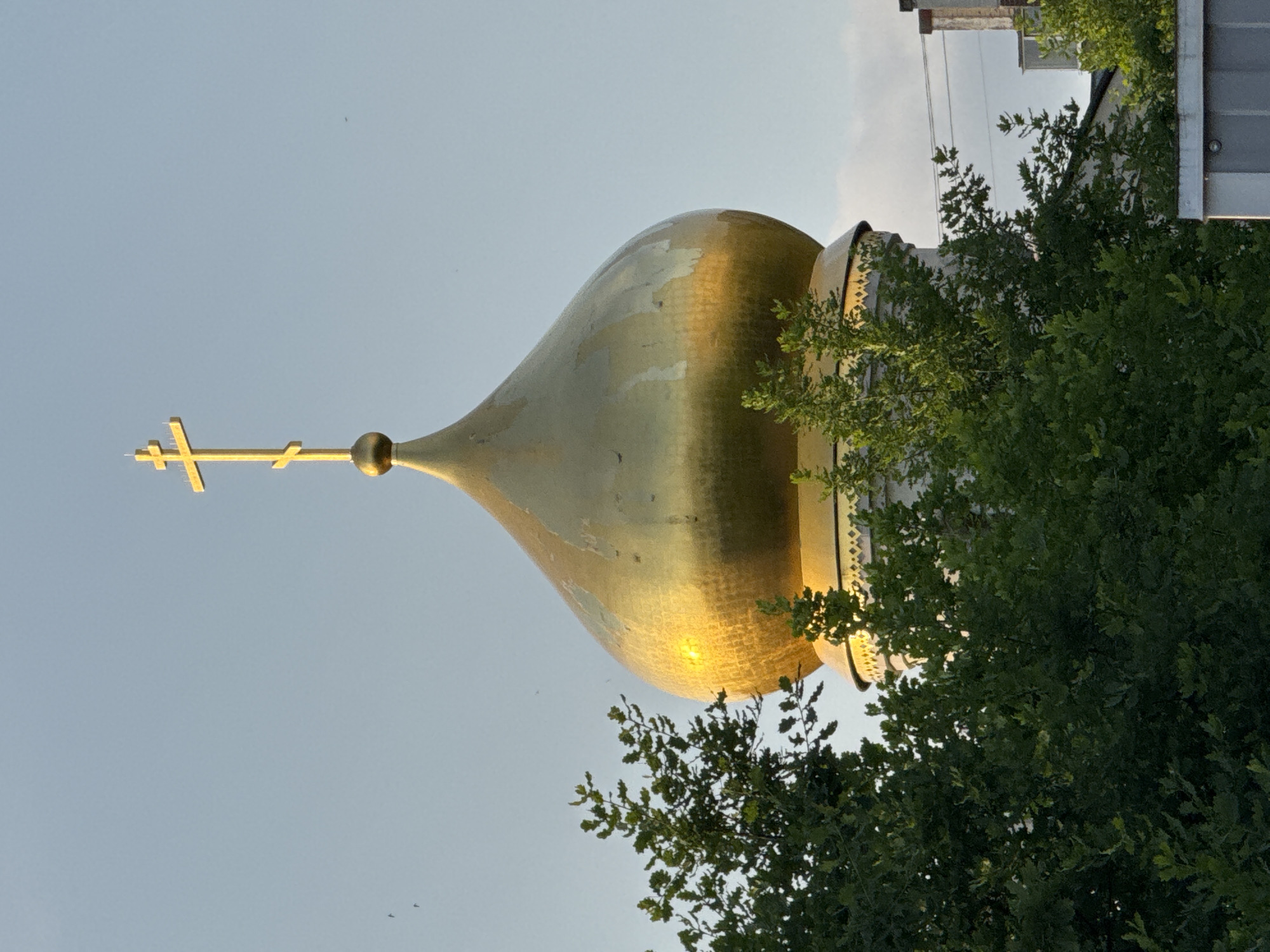An Orthodox Christian church dome, made of gold-plated material, is visible against a pale blue sky. The dome is topped with a golden Orthodox cross. Lush green trees partially obscure the view of the dome and the church structure below, which appears to be made of white stone with decorative patterns. The scene is captured during daylight, with sunlight reflecting brightly off the dome. The setting suggests an outdoor environment, possibly in the city of Kirishi, Russia, where such religious architecture is common. There are no people or animals visible in the image.