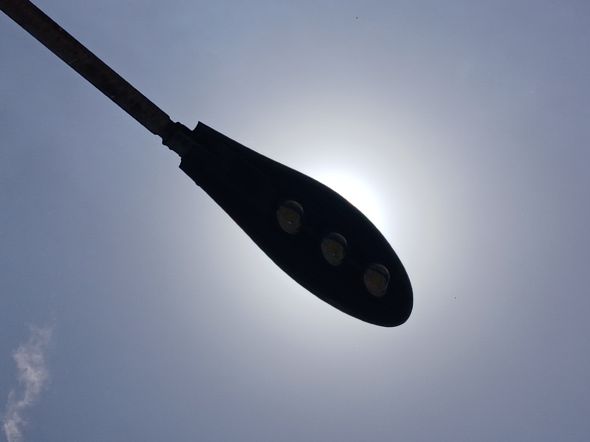 A street light fixture and its pole are silhouetted against a bright, hazy daytime sky. The dark, angled pole extends from the top-left towards the center, connecting to a modern, dark-colored LED street light fixture. The fixture is an elongated, flattened teardrop shape, with three circular LED components visible on its underside, appearing as lighter circles within the dark silhouette.
A brilliant, diffuse light source, presumably the sun, is positioned behind the street light fixture, creating a strong backlight that brightens the sky around it. The sky itself is a light blue-grey, suggesting either an overcast day or atmospheric haze scattering the sunlight. A small, wispy white cloud formation is visible in the bottom-left corner of the frame. The overall scene is static, capturing an inanimate object against the sky. No people are present, and no specific location details are visible within the image itself, though the context places it in Ciudad Guayana, Venezuela. There is no visible text.