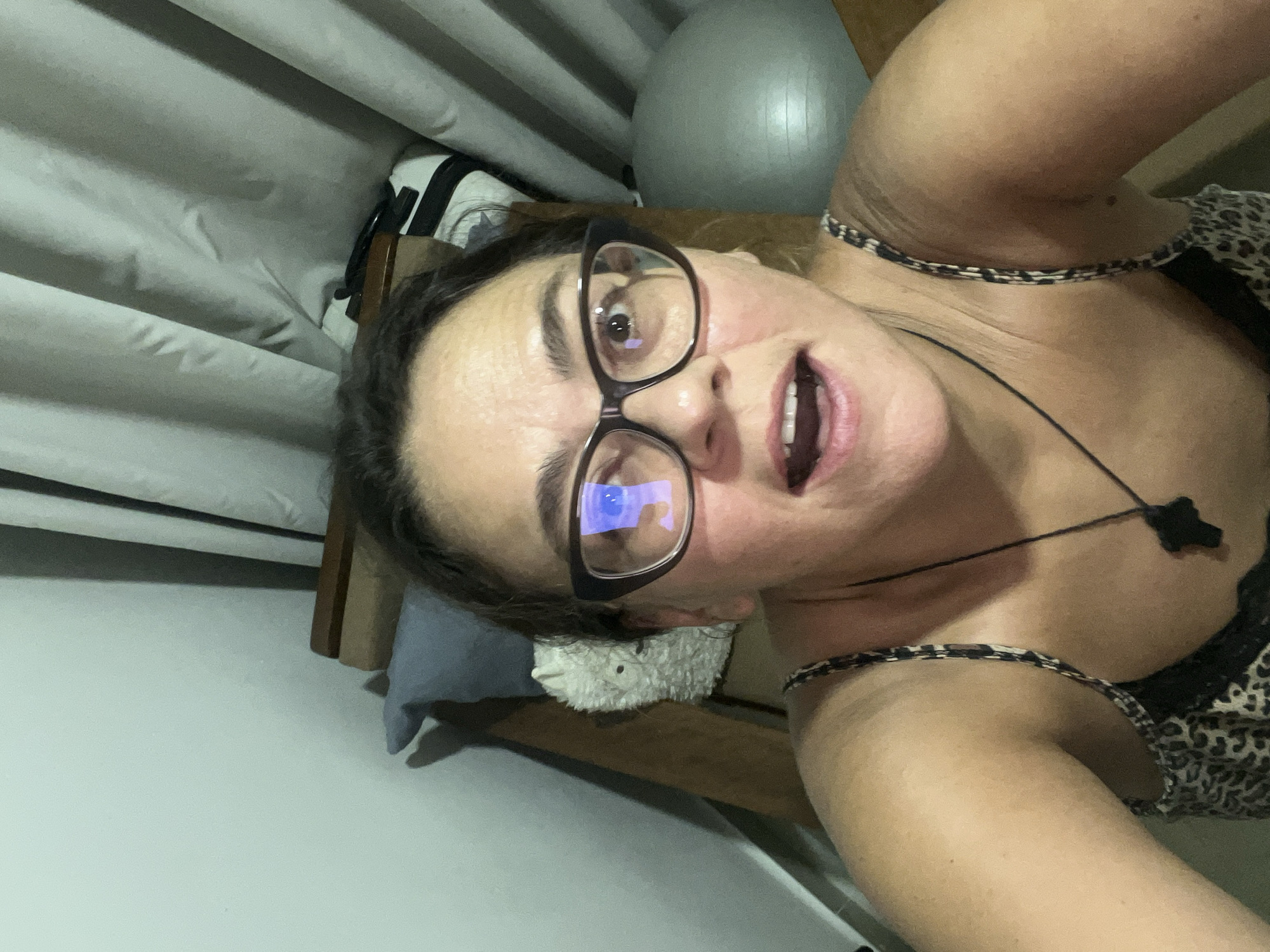 A woman with dark hair and glasses is taking a selfie indoors. She is wearing a leopard-print tank top. Behind her, there are light-colored curtains, a wooden structure, a pillow, a fluffy stuffed animal, and a grey exercise ball. The lighting suggests it is daytime. The location is not identifiable as Nha Trang Ward, Vietnam.