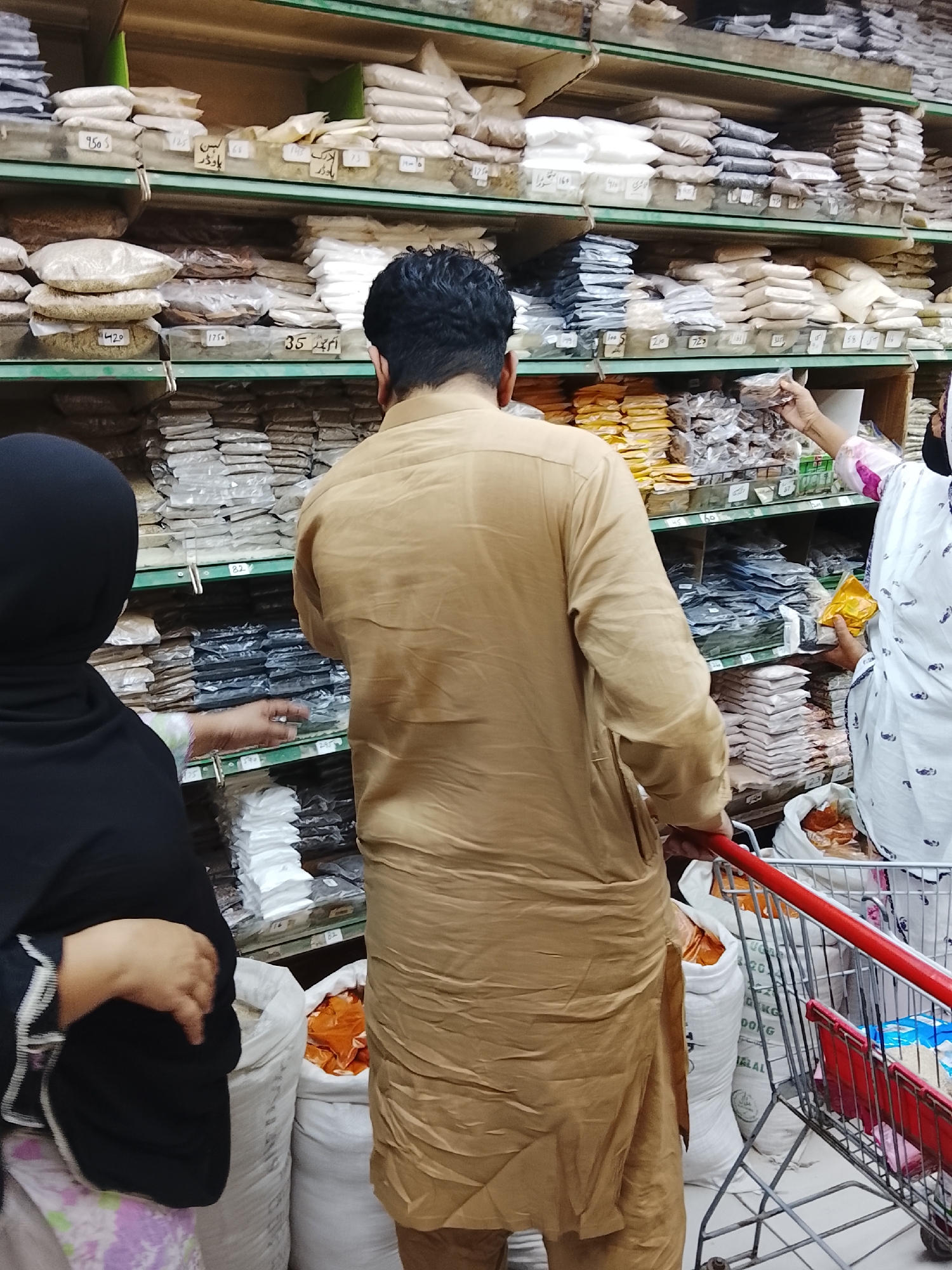 The image shows the interior of a store in Gulgasht Colony, Pakistan, featuring shelves stocked with bags and packages of goods, likely spices or grains. The goods are arranged on multiple shelves with price tags in Urdu or Arabic.
Three individuals are present: two are near the left side of the image, with one wearing a black hijab and the other in a brown tunic. The third person stands near the right side and is wearing a white and pink patterned garment and a face mask. A shopping cart, containing several white bags filled with items, is positioned near them.
The focus of the scene is on the shelves filled with a variety of products, and the individuals appear to be shopping for these items. The setting suggests a local grocery or spice store within the Gulgasht Colony area of Pakistan.