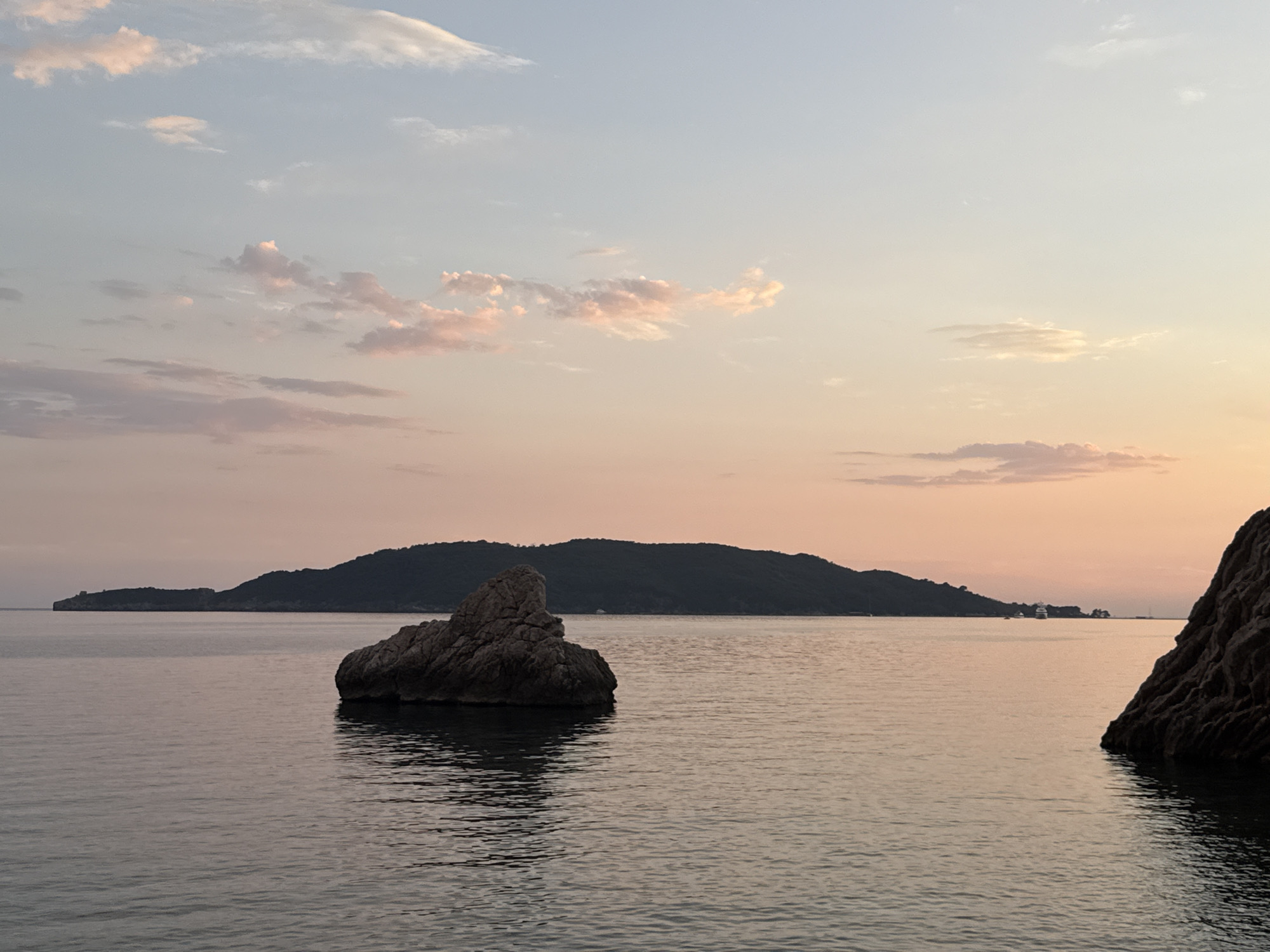 This media file depicts a serene seascape at dusk in Rafailovići, Montenegro. The scene is dominated by the calm, reflective water of the sea, bathed in the soft, pastel hues of the setting sun. In the foreground, a prominent, jagged rock formation emerges from the water on the right side of the frame, its dark silhouette contrasting with the lighter tones of the sea. Slightly to the left of the center, a smaller, similarly textured rock stands solitary in the water, casting a faint shadow.
In the distance, a lush, dark green, mountainous island forms the horizon. The island is gently sloped and covered in vegetation, suggesting a natural, undeveloped landscape. A few small boats or structures are faintly visible on the distant coastline of the island, hinting at human presence.
The sky above is a gradient of soft colors, transitioning from a pale blue at the top to a warm, rosy pink and orange near the horizon. Scattered, wispy clouds, tinged with pink, drift across the sky, adding texture and depth to the scene. The overall mood is peaceful and tranquil, evoking a sense of calm and the beauty of nature at the end of the day. The time of day is clearly sunset or twilight, with the soft lighting and muted colors characteristic of this period. No text is visible in the image.