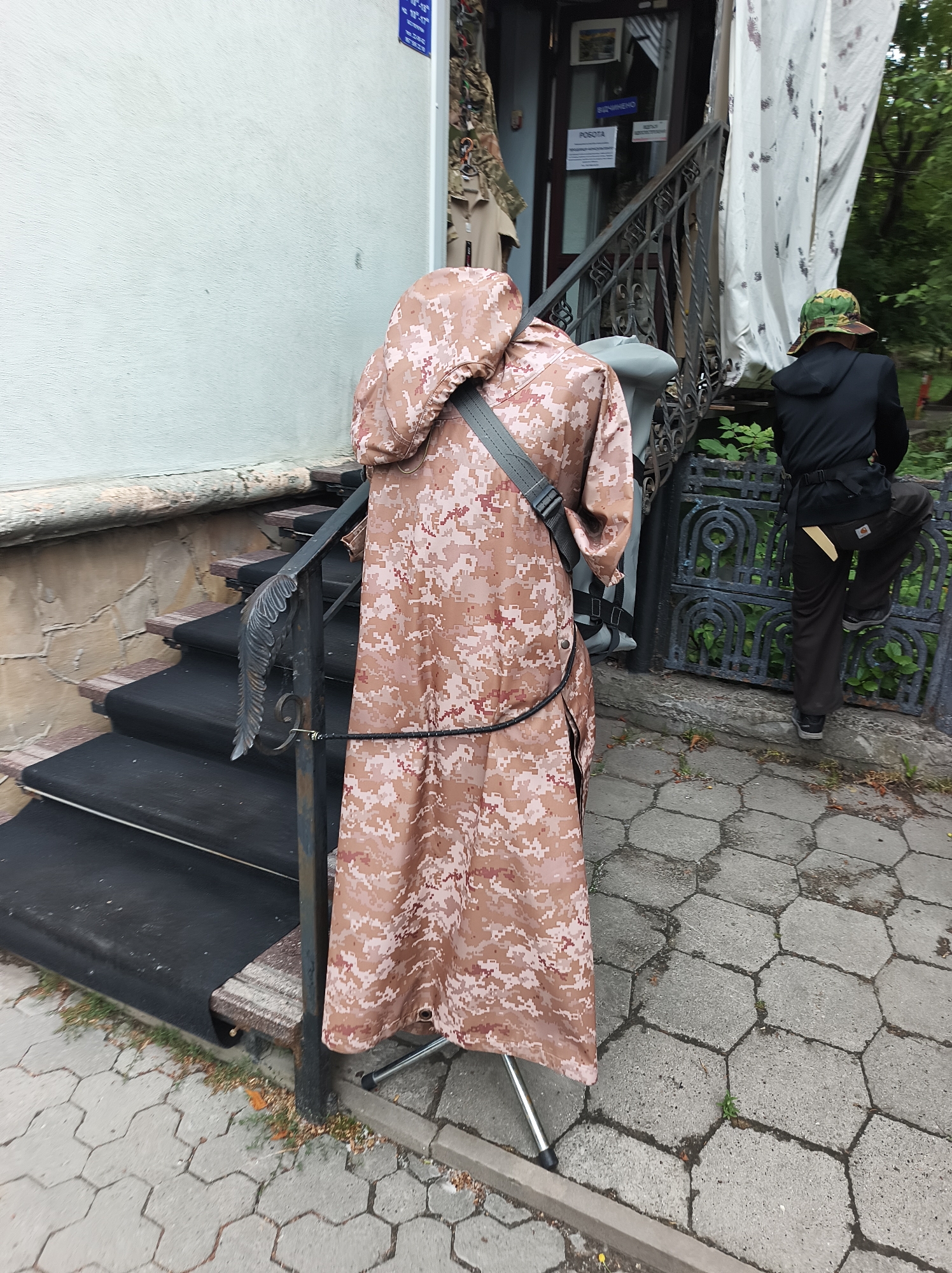 This image depicts an outdoor scene in Ternopil, Ukraine, featuring a retail entrance, a displayed garment, and an individual.
In the foreground, a sidewalk is paved with hexagonal grey concrete slabs. On the left, a metal display stand supports a long, hooded garment with a digital desert camouflage pattern, primarily in shades of tan, brown, and light grey. A grey strap diagonally crosses the upper back of the garment, and a thin black cord is tied around its midsection, extending towards the right.
Immediately behind the displayed garment, a set of approximately three to four stairs ascends to an entrance. These stairs are covered with a dark, possibly black, matting. A dark metal handrail with decorative, wing-like scrollwork on its lower section runs alongside the stairs. The building wall on the left is light-colored and textured, with a darker stone or stucco base beneath it.
At the top of the stairs is a dark-framed glass door, serving as the entrance. Visible on or through the door are several signs in Ukrainian: a white sign with "РОБОТА" (Work/Job) in black text, another white sign likely indicating "ВІДЧИНЕНО" (Open), and at least two blue signs with white text displaying operating hours, such as "10-19" and "10-17".
To the right of the stairs and entrance, a person is observed from the rear, partially obscured by a dark metal fence or gate. This individual wears a black hooded jacket or sweatshirt, dark brown or black pants, and a camouflage bucket hat with green and brown patterns. Their left foot rests on the low, decorative metal gate, which features repetitive arch-shaped motifs. A yellow or tan patch or strap is visible on the left thigh area of their pants. Behind the gate and the person, a white fabric sheet with dark, irregular patterns hangs, possibly a sunshade or curtain. Further in the background, green foliage, indicative of trees or bushes, is visible.