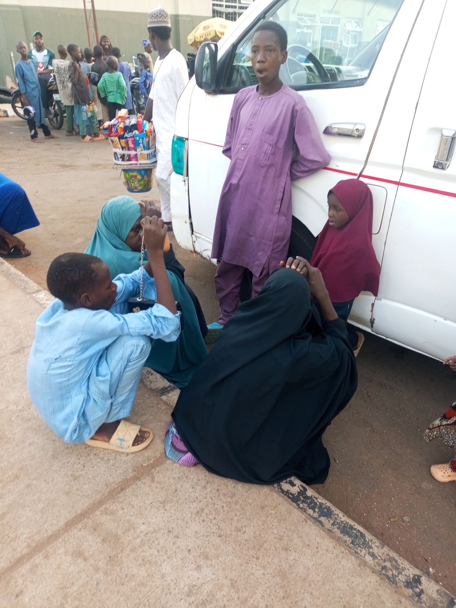 In Gombe, Nigeria, a group of individuals is gathered beside a white vehicle. A boy in a blue garment squats on the concrete ground, holding up a chain-like object. An adult in a green hijab sits beside him, while another adult in a dark-colored garment is bent over. Adjacent to the vehicle, a young boy in purple attire stands next to a girl wearing a maroon hijab. In the background, a vendor with a basket of snacks stands among a group of people. A motorcycle is also visible in the background. The scene appears to be outdoors, on a concrete surface near a building.
