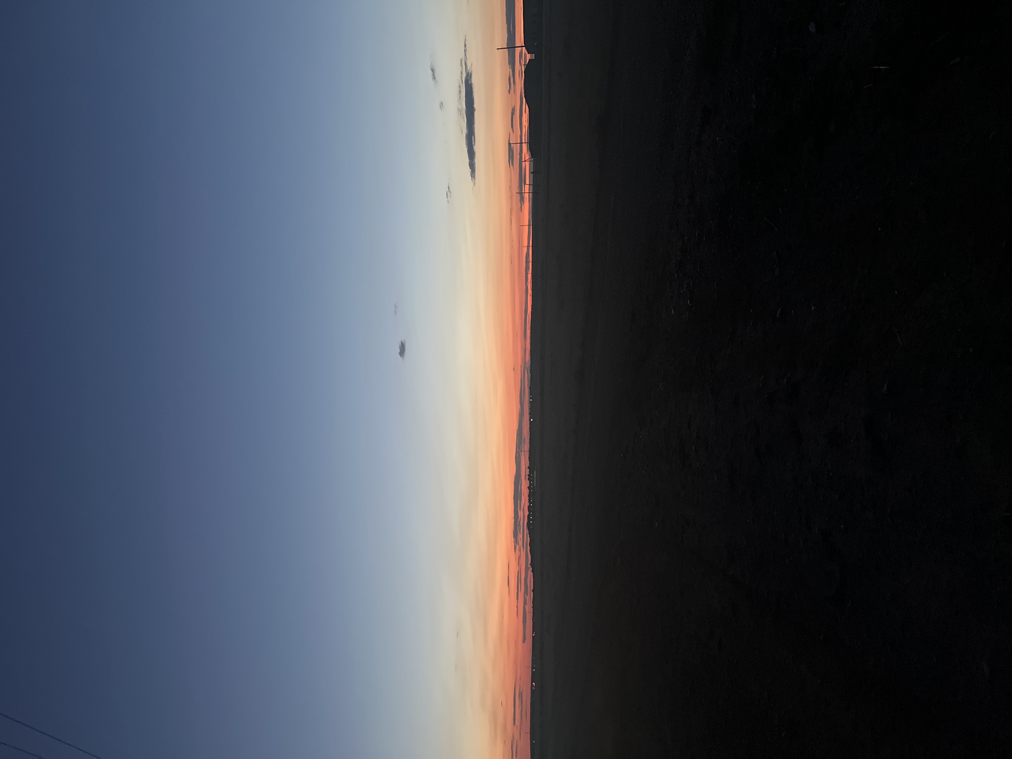 This is a vertical, eye-level, medium shot of a rural landscape at dusk in Arshan-zelmen, Russia. The sky is a gradient of deep blue at the top, transitioning to a vibrant orange and pink band just above the horizon, with thin, dark clouds scattered across it. The foreground is dominated by dark, barren earth, possibly a dirt road or field, with subtle variations in texture. In the middle ground, a faint line of structures and utility poles stretches across the horizon, silhouetted against the colorful sky. The overall impression is one of vastness and quiet solitude.