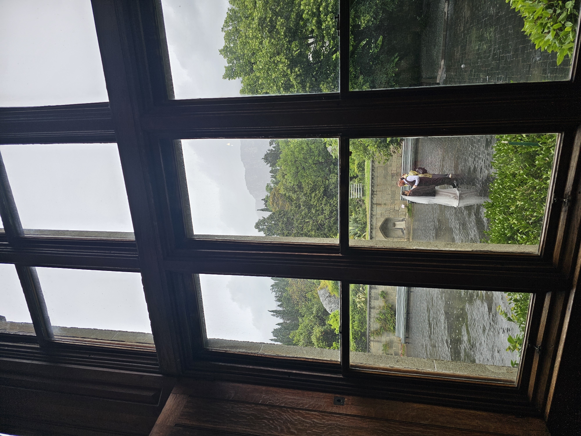 View from a window in Алупкинский городской совет, Ukraine, showing a couple standing on a stone bridge in what appears to be a park or garden. The trees and sky are overcast, suggesting a rainy day. The architectural style visible through the window includes traditional woodwork.