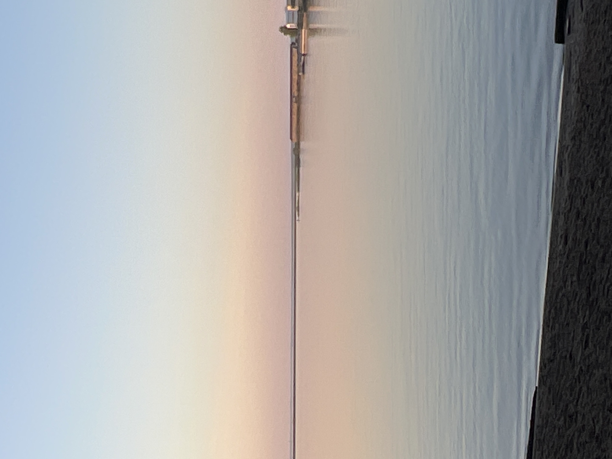 A long pier stretches into a body of water, likely a lake or river, under a pastel-colored sky, probably at sunrise or sunset. The sky transitions from light blue to a warm, pale orange. The pier appears to be constructed of metal and wood. The water's surface is relatively calm. To the right of the water is a dark, textured surface, potentially land or a pier surface. I don't have enough information to place this image in the city of Chaikovsky, Russia.