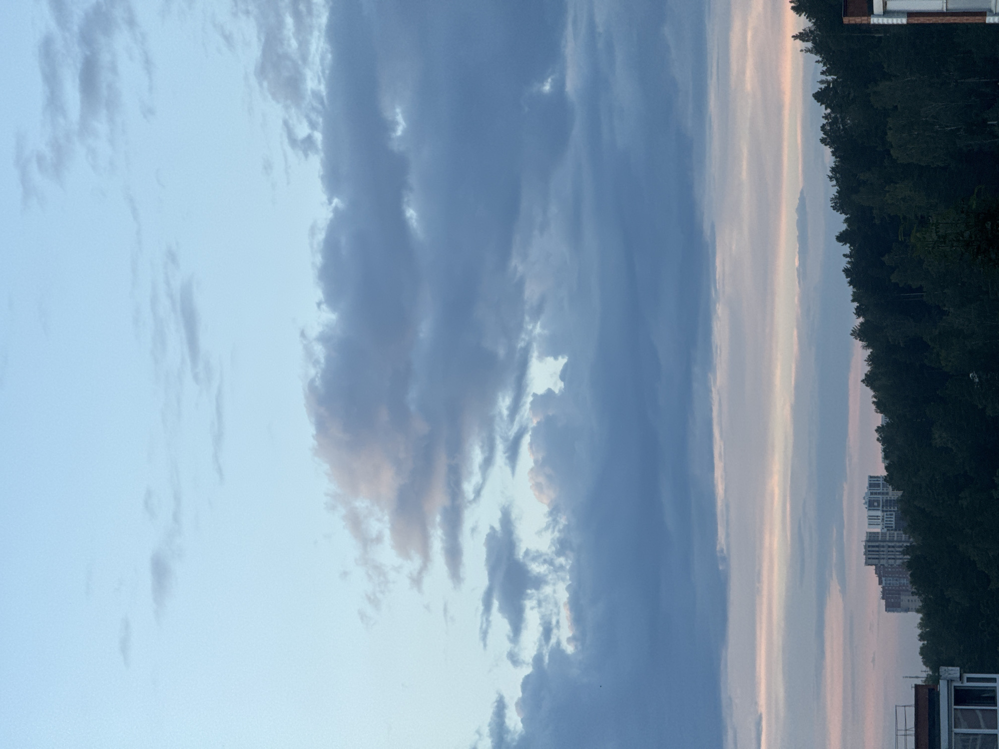 The image depicts a wide-angle view of a serene sky above a cityscape at dusk. The sky transitions from a pale blue at the top to hues of pink and grey near the horizon, with scattered clouds in various shades of blue and white. In the middle ground, a dense forest of dark green trees forms a natural barrier. Beyond the trees, a cluster of tall, modern residential buildings, characterized by their white and beige facades and numerous windows, are visible. The lower foreground shows the edge of a building with a red-tiled roof, and a part of a white structure with windows. The scene appears to be an outdoor urban landscape in Markova, Russia, captured during the twilight hours.