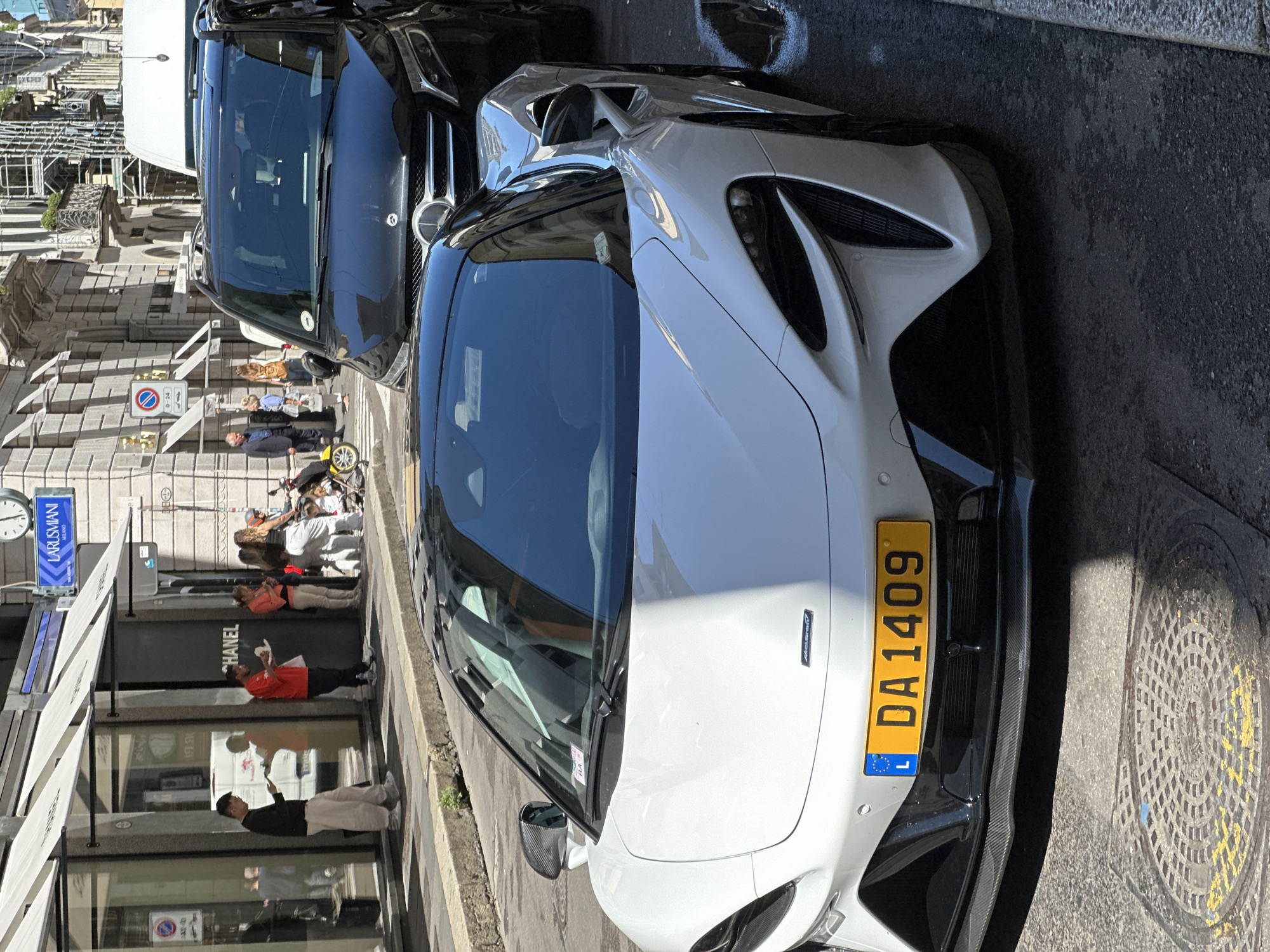 An eye-level shot captures a bright white McLaren supercar parked on an urban street, possibly in Milan, Italy, given the context. The McLaren, likely a 720S or similar model, features sleek lines and carbon fiber elements on its front bumper. Its front-mounted license plate is yellow with black characters, displaying "DA 1409" and a small "L" for Luxembourg.
Behind the McLaren, a dark-colored Mercedes-Benz SUV or large sedan is also parked. The street appears to be paved with asphalt, with some ornate manhole covers visible on the ground.
The background shows a bustling street scene with several pedestrians. People are walking, standing, and interacting near storefronts. One shop clearly displays the "CHANEL" logo on its dark facade, with white awnings above. Another blue sign further down the street reads "LARUSMIANI MILANO." A circular Italian street sign, indicating "no parking" or a specific restriction, is also visible with "0-24" written on it.
The lighting suggests a sunny day, with clear shadows cast by the vehicles and buildings, indicating midday or early afternoon. The overall atmosphere is that of a lively city street.