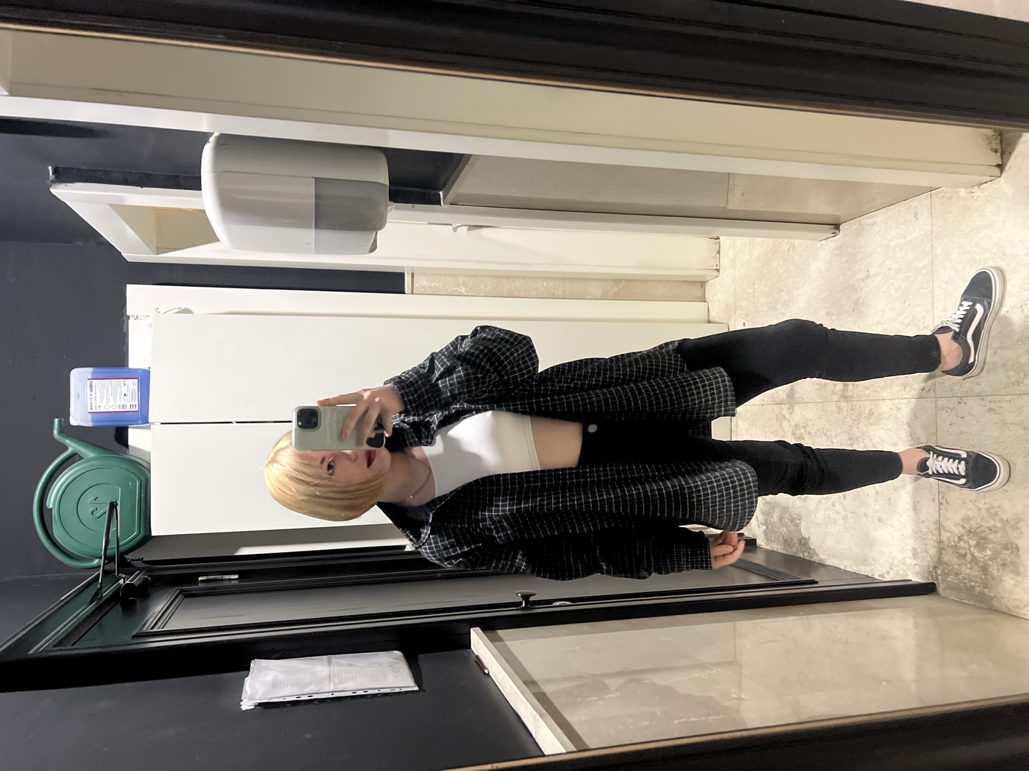 A young woman is standing in a restroom, taking a selfie in the mirror. She is wearing black leggings, a white crop top, and a black and white plaid shirt. She has short blonde hair and is holding a smartphone in her right hand. The restroom has tiled floors and walls. A paper towel dispenser is mounted on the wall, and a green hose reel is attached to the door. The image was taken in Budva, Montenegro.