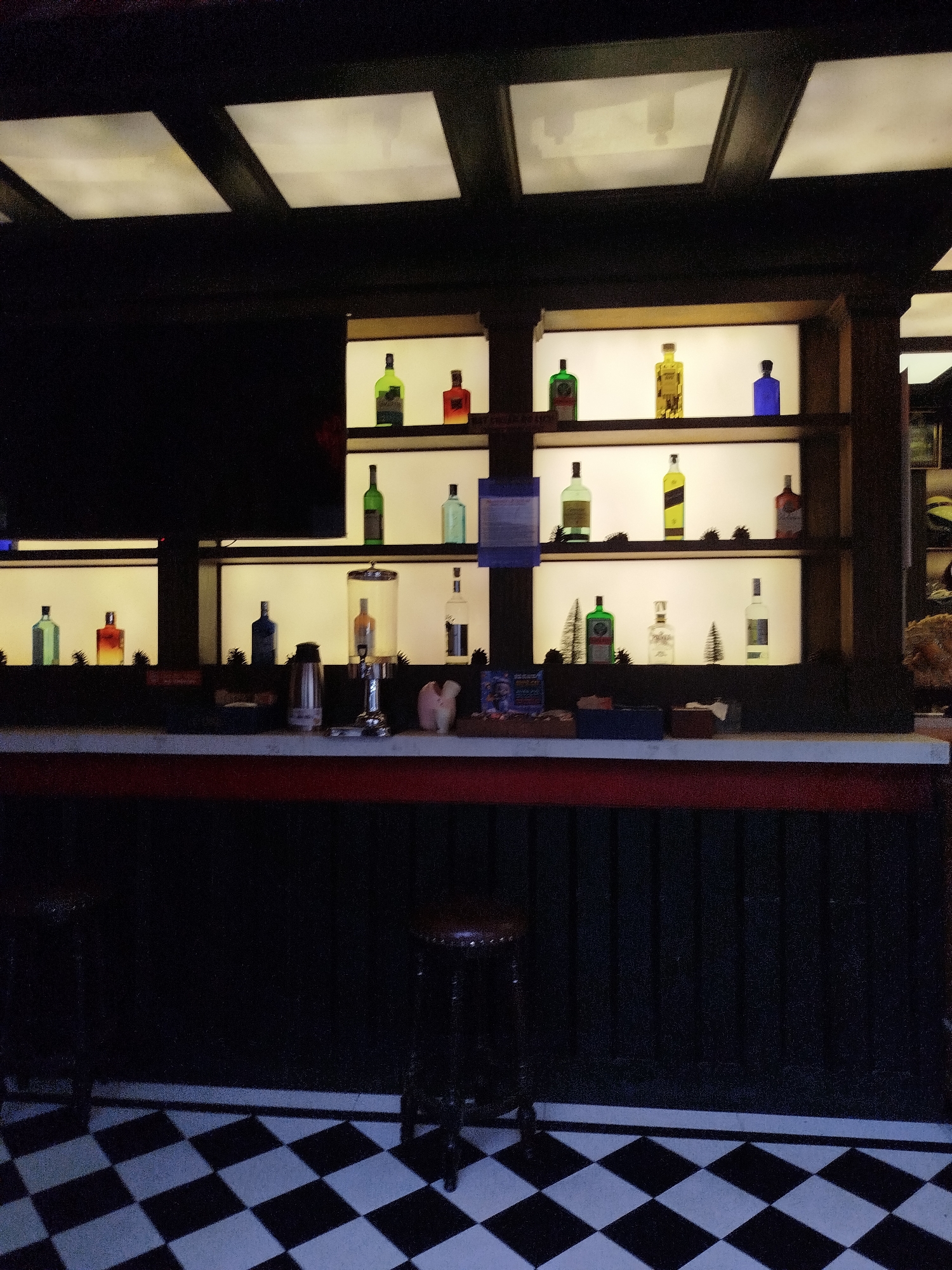 The image shows the interior of a bar. The bar counter is in the foreground with two bar stools in front. Behind the bar is a shelving unit displaying various bottles of alcohol, illuminated from behind. The ceiling has a grid pattern with recessed lighting. The floor is tiled with a black and white checkerboard pattern. The lighting is dim. The location is possibly Ho Chi Minh City, Vietnam.