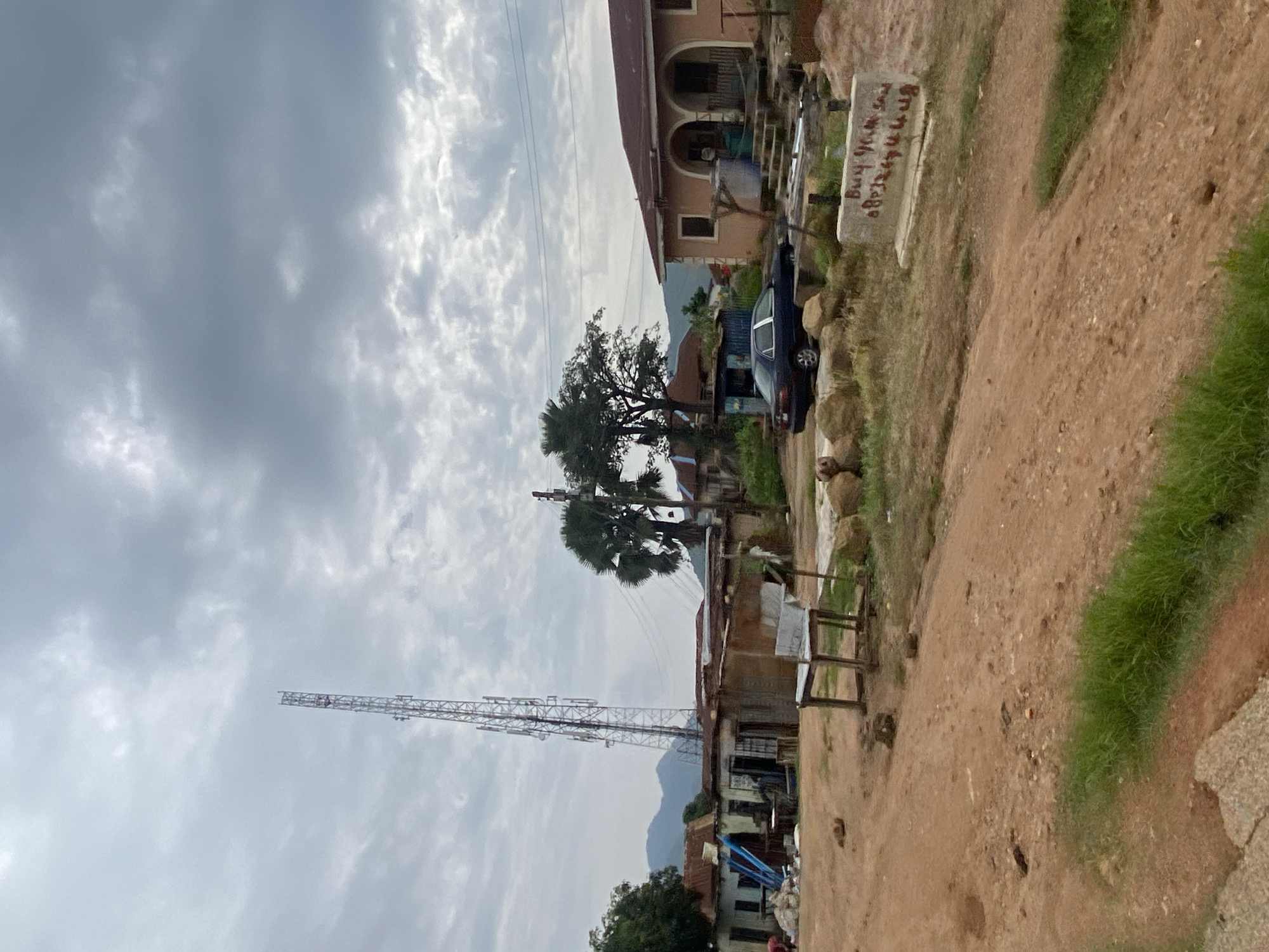 The image depicts a rural or semi-urban landscape in Jos, Nigeria, characterized by a dirt road, simple structures, and natural elements under a cloudy sky. Dominating the left side of the frame is a tall, slender telecommunications tower. Adjacent to it are several modest, single-story buildings with corrugated metal roofs, appearing to be shops or residences. One of these structures has blue pipes leaning against its exterior. In the foreground, a weathered wooden bench or chair sits in the middle of the dirt road. To the right, a more substantial, light-colored house with arched doorways and a reddish-brown roof is visible. A dark-colored sedan is parked in front of this house. The landscape is dotted with trees, including palm trees, and in the background, rolling hills can be seen. A concrete block with red graffiti-like writing sits in the right foreground. The overall impression is one of a developing area, blending infrastructure with a natural, somewhat rugged environment.