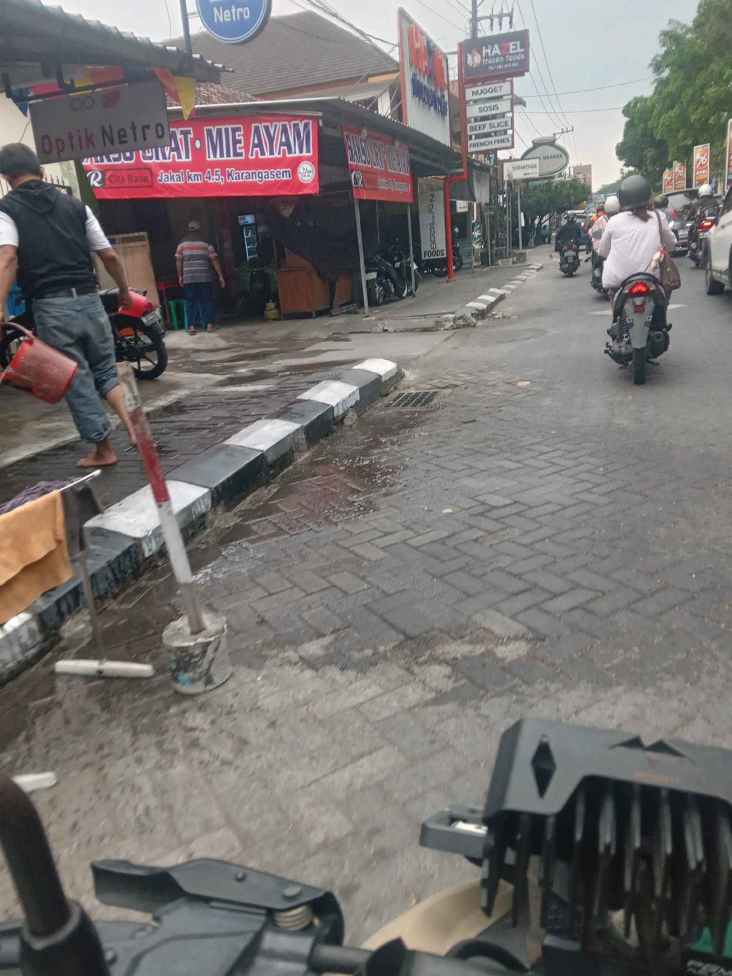 The image depicts a street scene in Depok, Indonesia, likely on a cloudy day, with a focus on the storefronts and traffic. In the foreground, a person wearing jeans and a black vest is visible, carrying a red bucket. To the left, a red motorcycle is parked. Directly behind the person, a sign for "Optik Netro" is present. Further down the street, a prominent red banner displays "MI AM AYAM" in large white letters, with "Jakal km 4.5, Karangasem" below it, indicating the location. Another sign reads "HAZEL frozen foods" with a list of items including "NUGGET", "SOSIS", "BEEF SLICE", and "FRENCH FRIES". The street itself is paved with interlocking bricks, and the area appears wet, suggesting recent rain. Several people are seen on motorcycles, navigating the road, with one rider in a white shirt and helmet prominently featured in the middle ground. The overall environment suggests a bustling commercial area with an outdoor market or food stalls.