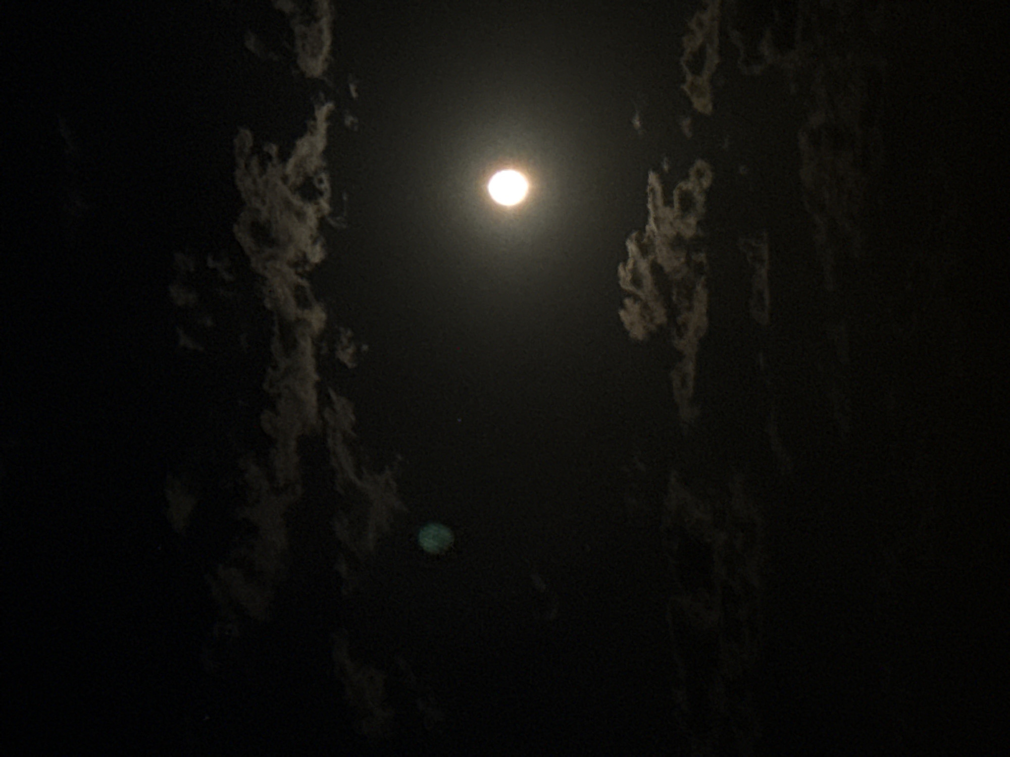 The image displays a nocturnal scene featuring a bright, circular celestial body, presumably the moon, centered horizontally in the upper portion of the frame. The moon appears as an intensely luminous, overexposed white disc, radiating a soft glow that subtly illuminates its immediate surroundings.
On both the left and right sides of the image, dark, irregularly shaped clouds are visible, stretching vertically. These clouds have a textured, wispy appearance and seem to be partially illuminated from behind or by the moon's light, creating a sense of depth against the dark sky. They frame the moon, suggesting a channel-like opening through the cloud cover.
A distinct, round, greenish-blue artifact, characteristic of a lens flare, is present in the lower-central area of the image, positioned slightly to the left. The overall setting is a cloudy night sky. No specific activities or events are depicted, nor is any text visible.