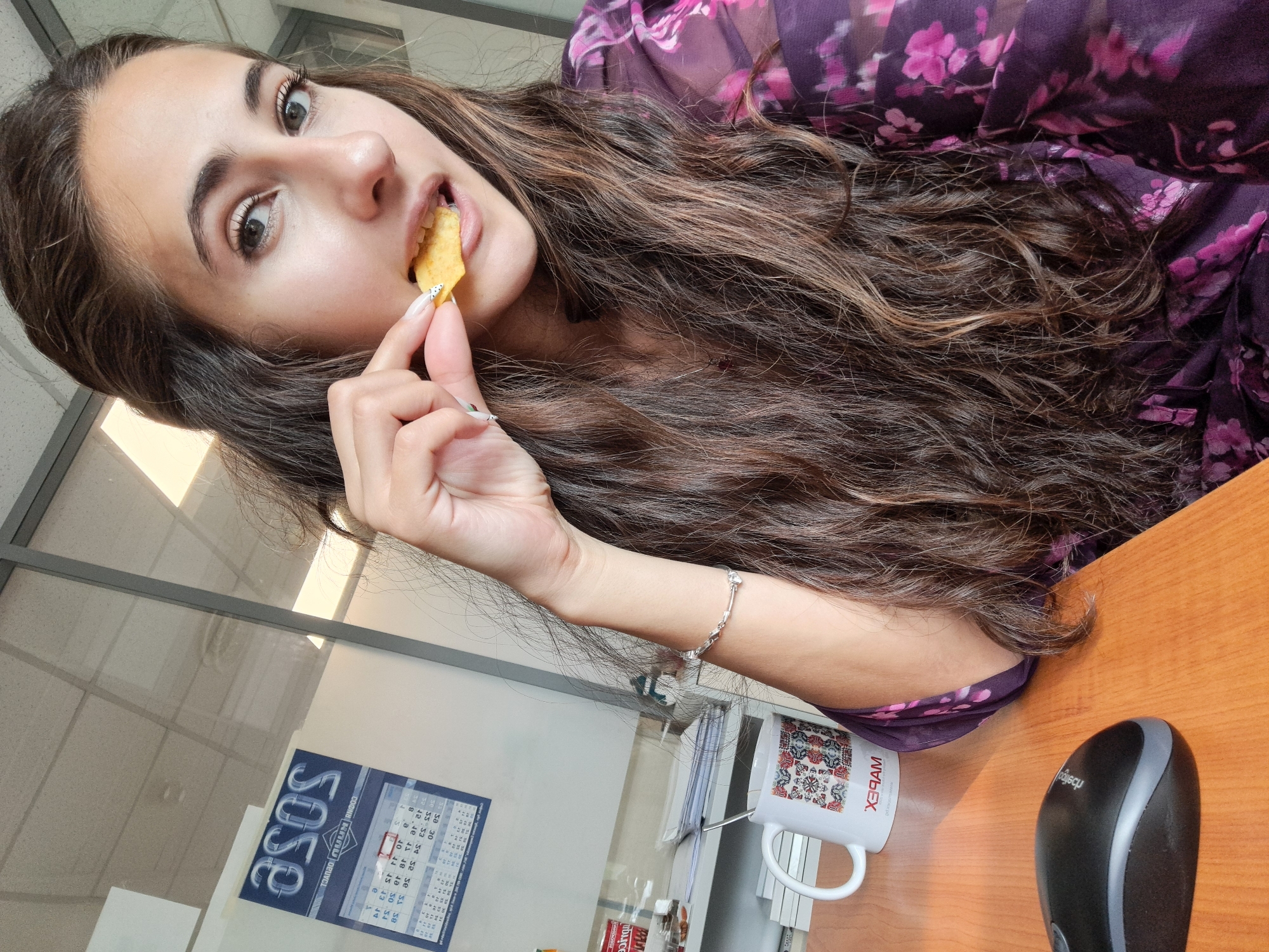The image shows a young woman captured in a candid, close-up shot, likely indoors in an office environment. Her long, dark, wavy hair cascades over her shoulders and chest. She is wearing a purple top with a floral pattern.
The woman is in the act of eating, holding a yellow, triangular chip to her mouth with her right hand. Her fingernails have a light polish with small black dot accents. She looks directly towards the viewer with a neutral, slightly focused expression. A delicate silver bracelet is visible on her left wrist.
The setting appears to be a workplace, with a light wooden desk in the foreground. On the desk, a white mug featuring a colorful, intricate pattern and the text "МАВЕХ" and "ХРАМ" (HRAM, meaning 'temple' in Bulgarian) is visible. A black and grey computer mouse, with the partial brand name "logiq", rests nearby.
In the background, behind the woman, a calendar for "2023" is affixed to a wall or glass partition. Partially visible text includes the months "МАЙ" (May), "ЮНИ" (June), "ЮЛИ" (July), and "АВГУСТ" (August), along with "СОФИЯ". Bright daylight streams through windows or glass panels in the background, illuminating the space. The scene suggests a casual moment during the day in Sofia, Bulgaria.
