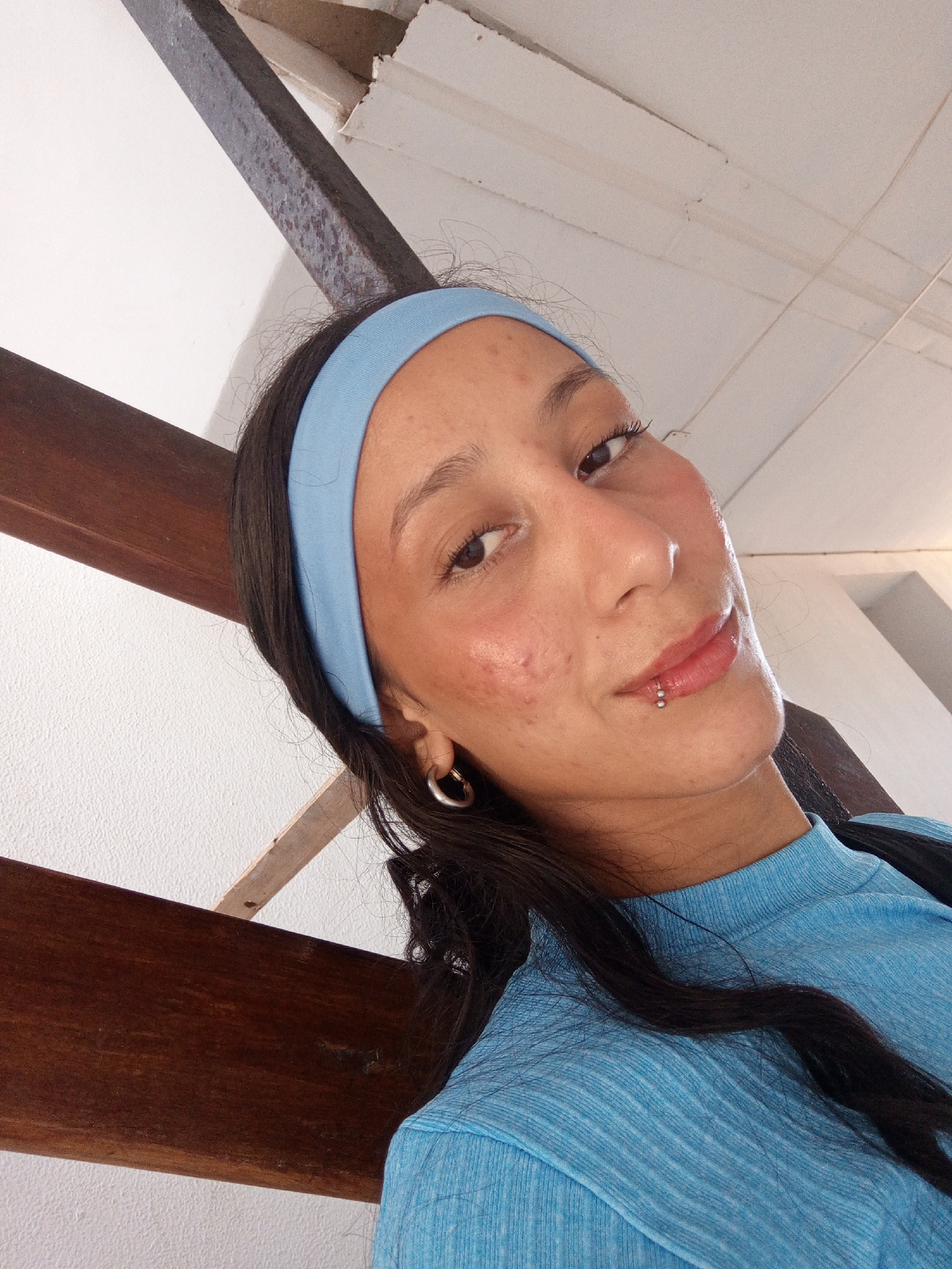 A young adult female poses for a selfie. She has dark hair and a light blue headband. She wears a matching light blue long-sleeved shirt. She has a lip piercing and wears hoop earrings. Her complexion shows some blemishes. The background suggests an interior location with white walls and wooden beams, possibly a staircase or architectural detail. The location in the city of Porlamar, Venezuela is not discernible from the image.