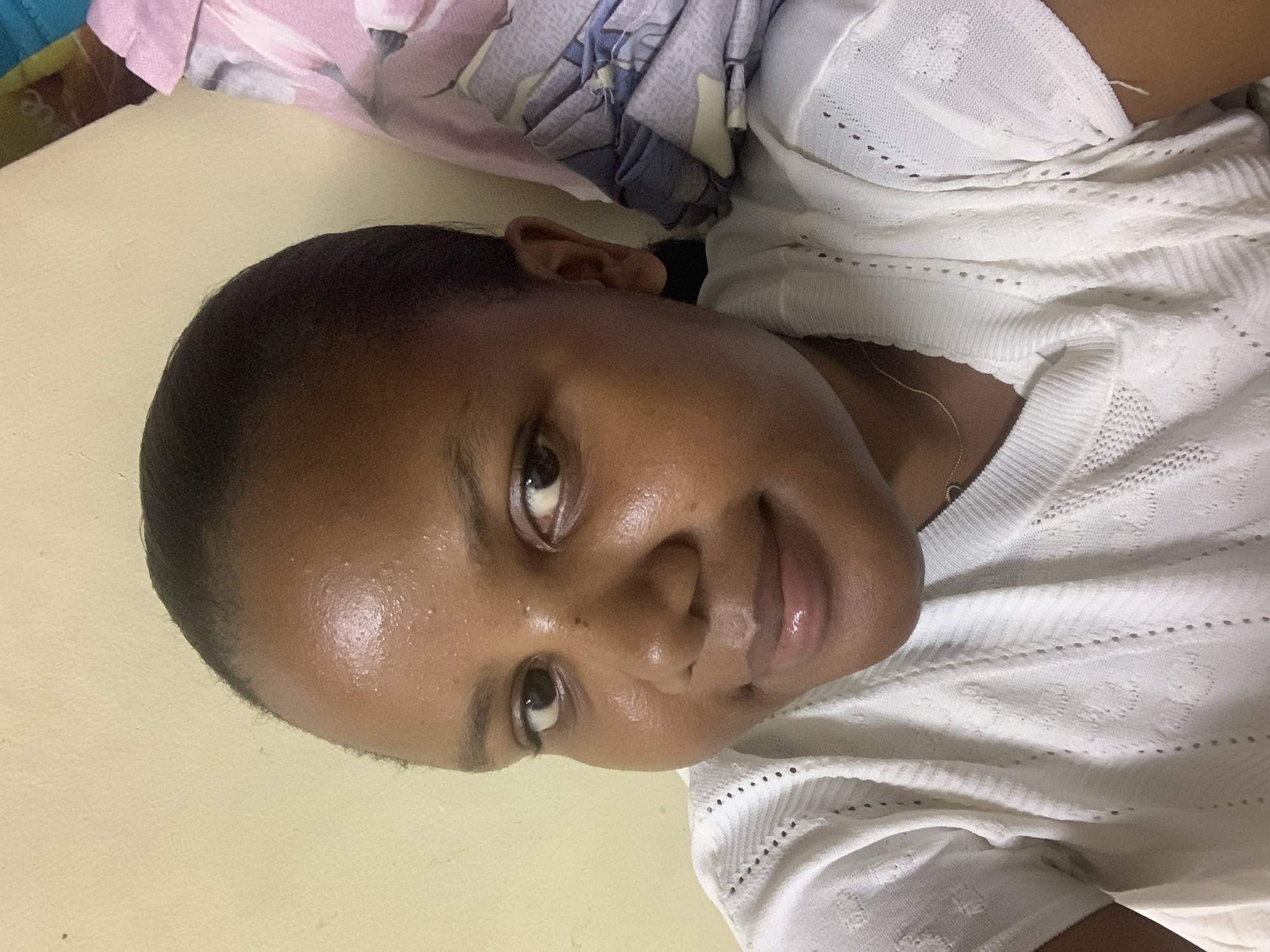 A medium shot depicts a young woman with dark skin, glossy lips, and dark eyes. She is looking slightly upwards and towards the viewer. She appears to be wearing a white, textured knit top. The background is a solid light yellow color with a patterned bedsheet visible in the top left corner. There is no information in the image to confirm if the location is Dar es-Salaam, Tanzania.